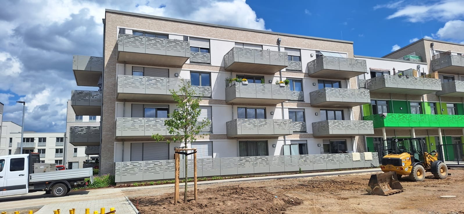 Nowoczesny budynek wielorodzinny z balkonami zabezpieczonymi ażurowymi balustradami w kolorze szarym, na pierwszym planie małe drzewko i maszyna budowlana.