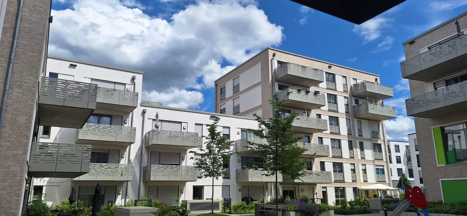 Nowoczesne budynki mieszkalne z balkonami o metalowych, ażurowych barierkach, widok z poziomu dziedzińca z zielenią i placem zabaw.