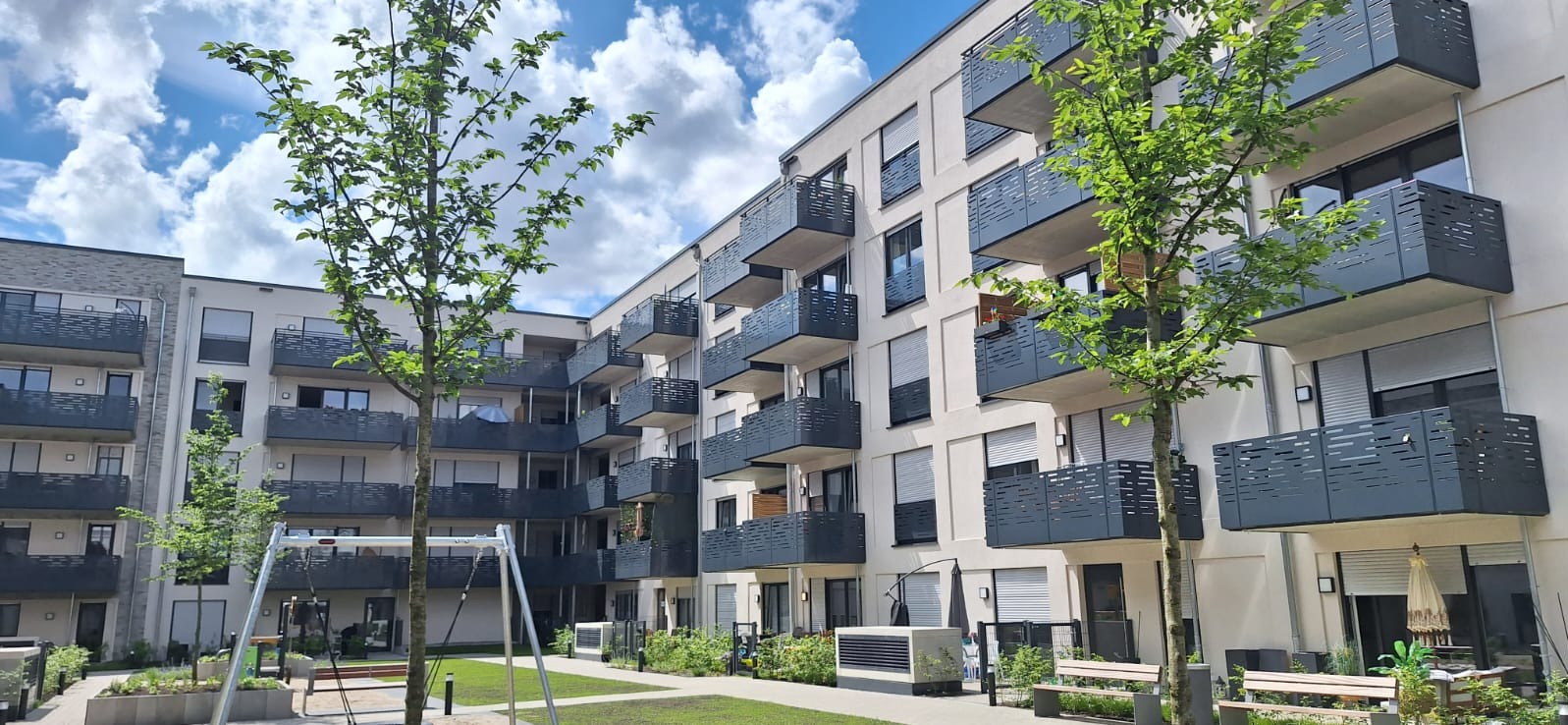 Elewacja budynku wielorodzinnego z balkonami zabezpieczonymi metalowymi balustradami o nowoczesnym wzorze, widok z dziedzińca z trawnikiem i drzewami.