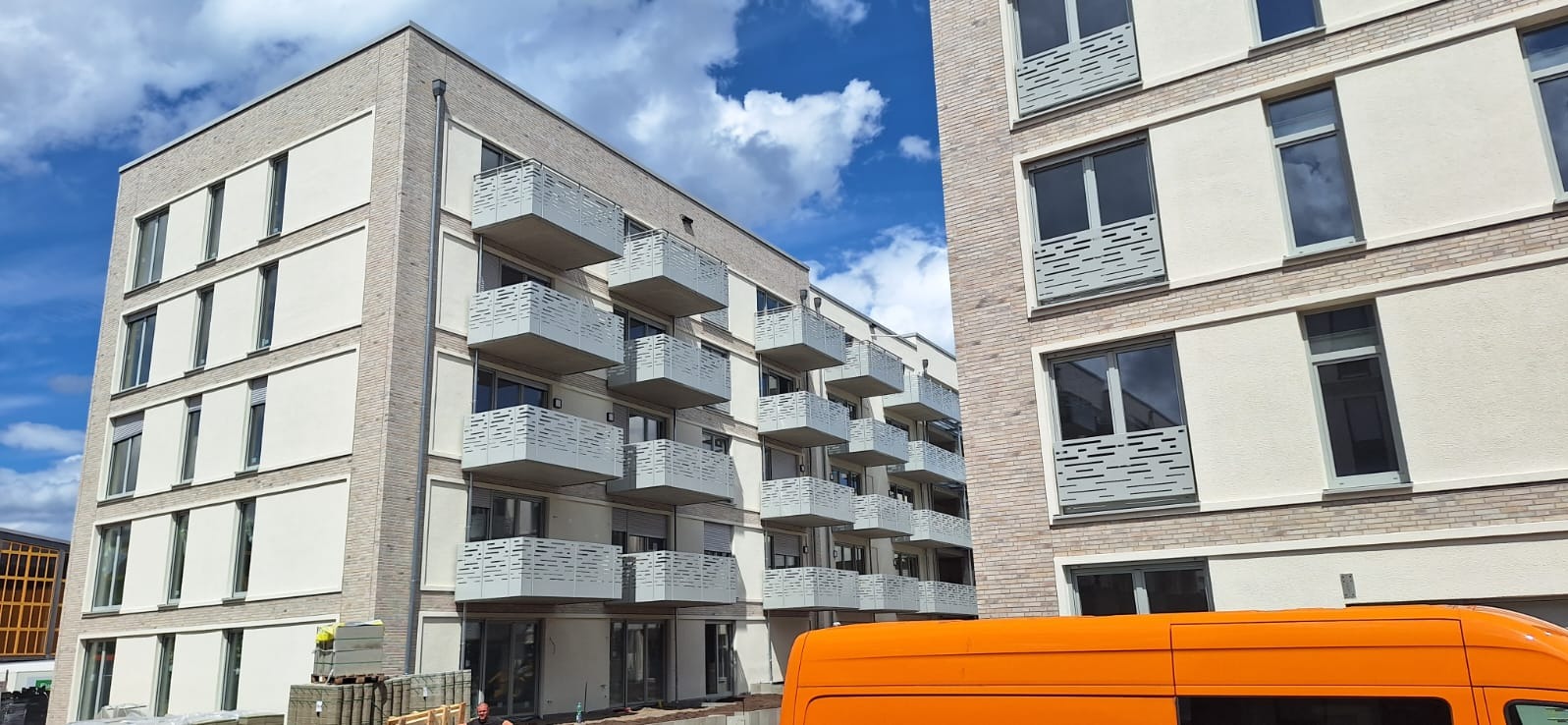 Nowoczesny budynek mieszkalny z balkonami zabezpieczonymi ażurowymi balustradami, widok z poziomu ulicy z fragmentem pomarańczowego samochodu dostawczego.