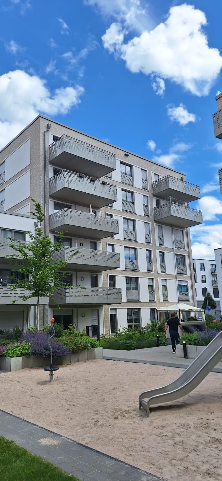 Nowoczesny budynek mieszkalny z balkonami z ażurowymi barierkami, widok z placu zabaw w słoneczny dzień.