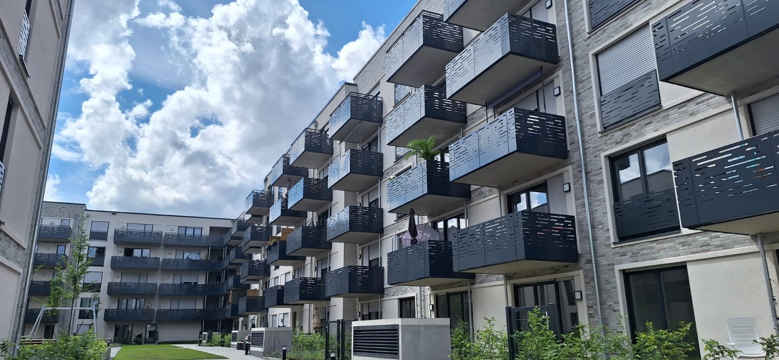 Elewacja budynku z licznymi balkonami zabezpieczonymi metalowymi balustradami o ażurowym wzorze, widok z perspektywy trawnika w słoneczny dzień.