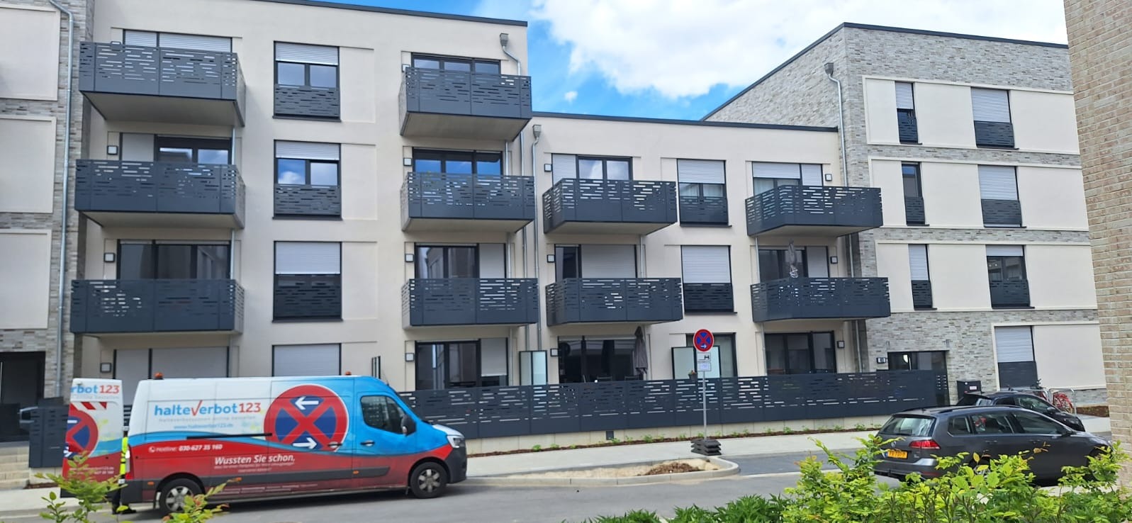 Nowoczesny budynek wielorodzinny z balkonami zabezpieczonymi szarymi, metalowymi balustradami o ażurowym wzorze, na pierwszym planie zaparkowany samochód dostawczy z niebiesko-czerwonymi grafikami.