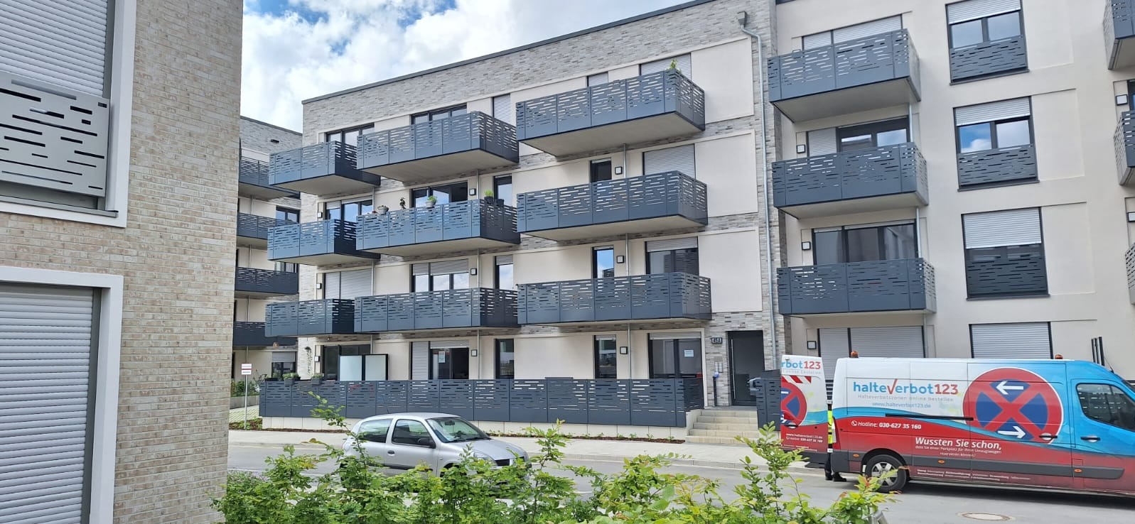 Nowoczesny budynek wielorodzinny z balkonami wyposażonymi w ażurowe, metalowe barierki w kolorze grafitowym, widok z poziomu ulicy, zieleń w tle, zaparkowany samochód i furgonetka.