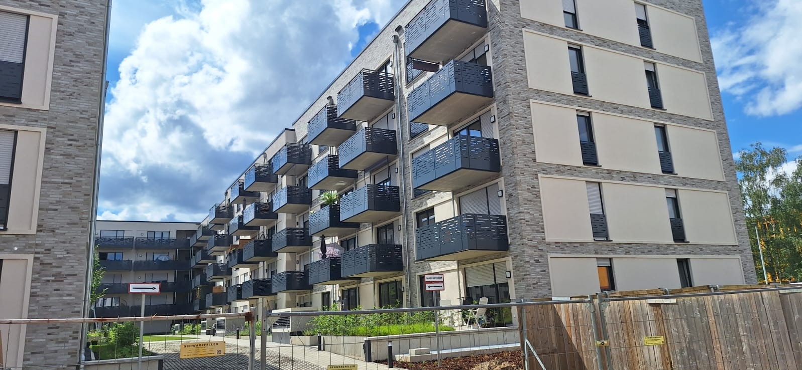 Nowoczesny budynek mieszkalny z balkonami z ażurowymi barierkami, widok z poziomu ulicy, częściowo zasłonięty ogrodzeniem budowlanym.