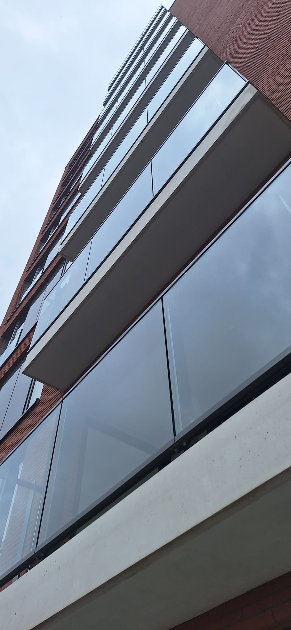 Nowoczesny budynek mieszkalny z balkonami zabezpieczonymi szklanymi balustradami na betonowych płytach, widziany z dołu pod kątem.