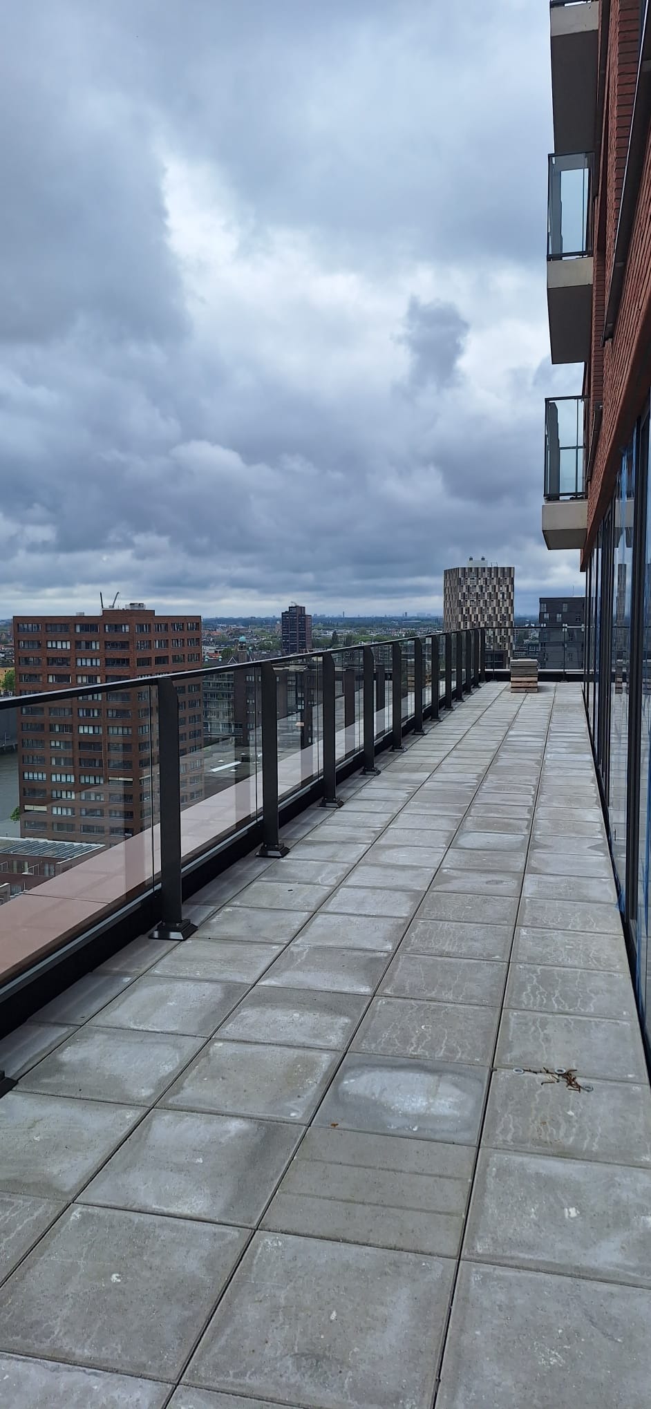 Długi balkon z szarymi płytkami i szklaną balustradą z czarnymi słupkami, widok na miasto w pochmurny dzień.
