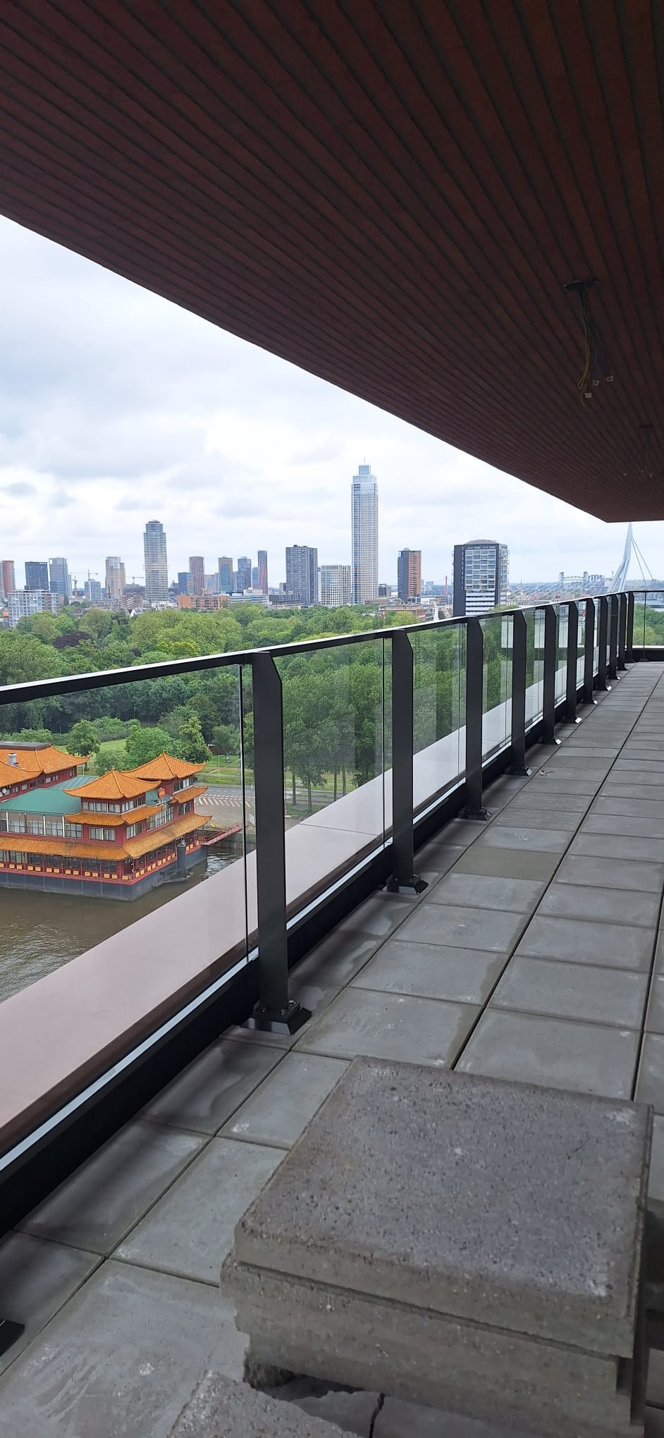 Szklana balustrada z czarnymi słupkami na tarasie z widokiem na panoramę miasta i pływającą restaurację w stylu orientalnym.