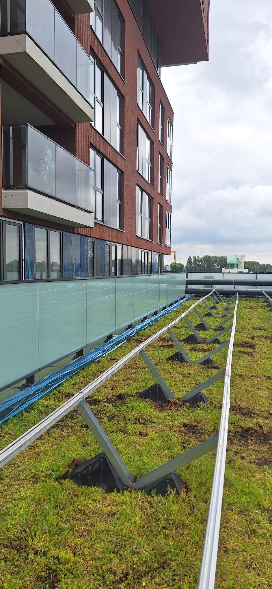 Montaż konstrukcji wsporczej na zielonym dachu nowoczesnego budynku z szklanymi balustradami balkonowymi.