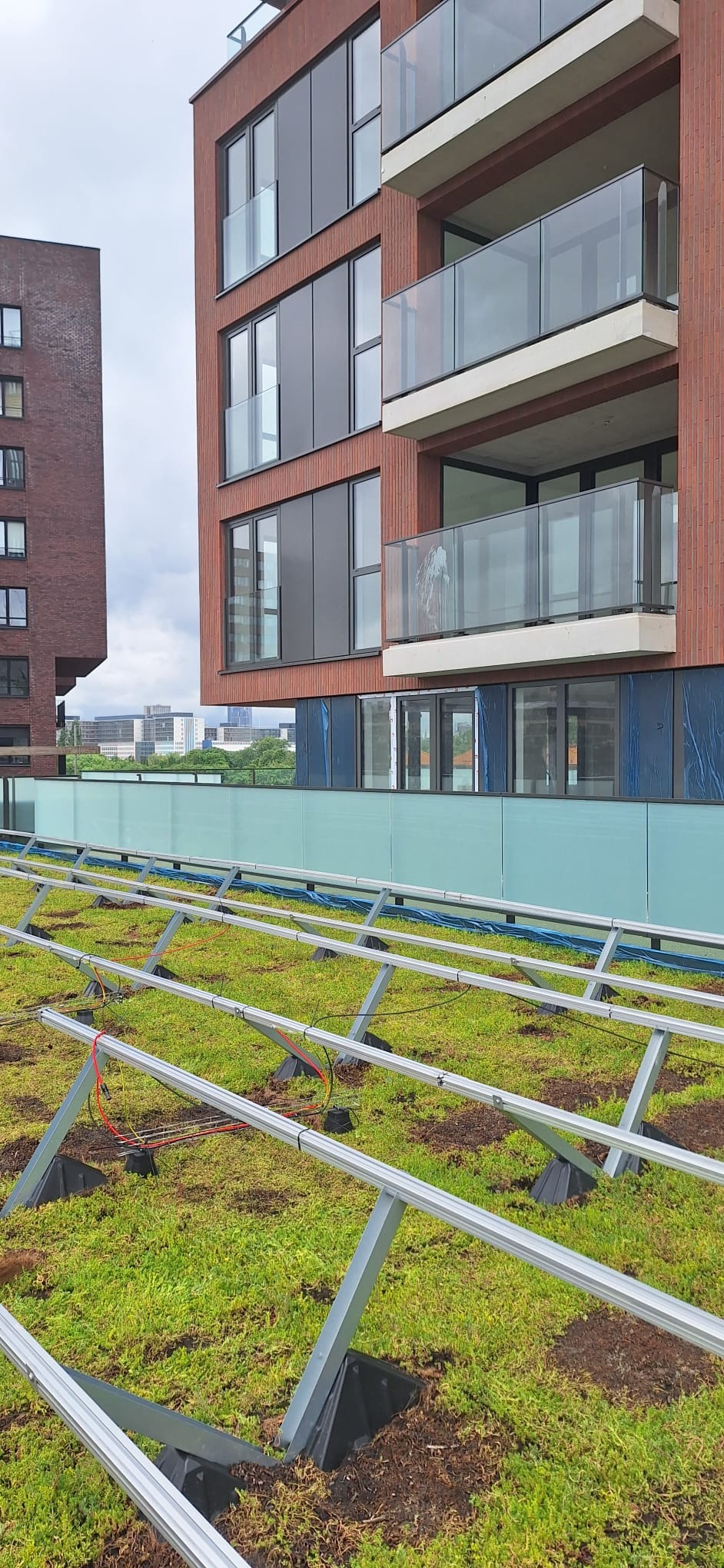 Montaż stelażu pod panele słoneczne na zielonym dachu budynku wielorodzinnego, widoczne balustrady balkonowe ze szkła.