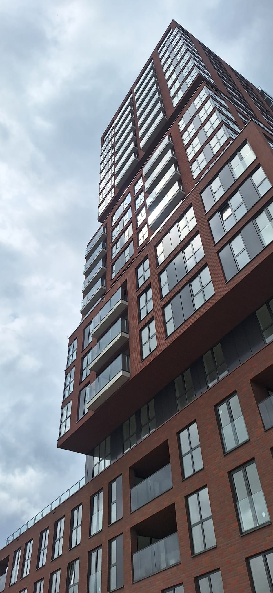 Wysoki budynek mieszkalny z cegły z balkonami z przezroczystymi panelami, widok z dołu w pochmurny dzień.