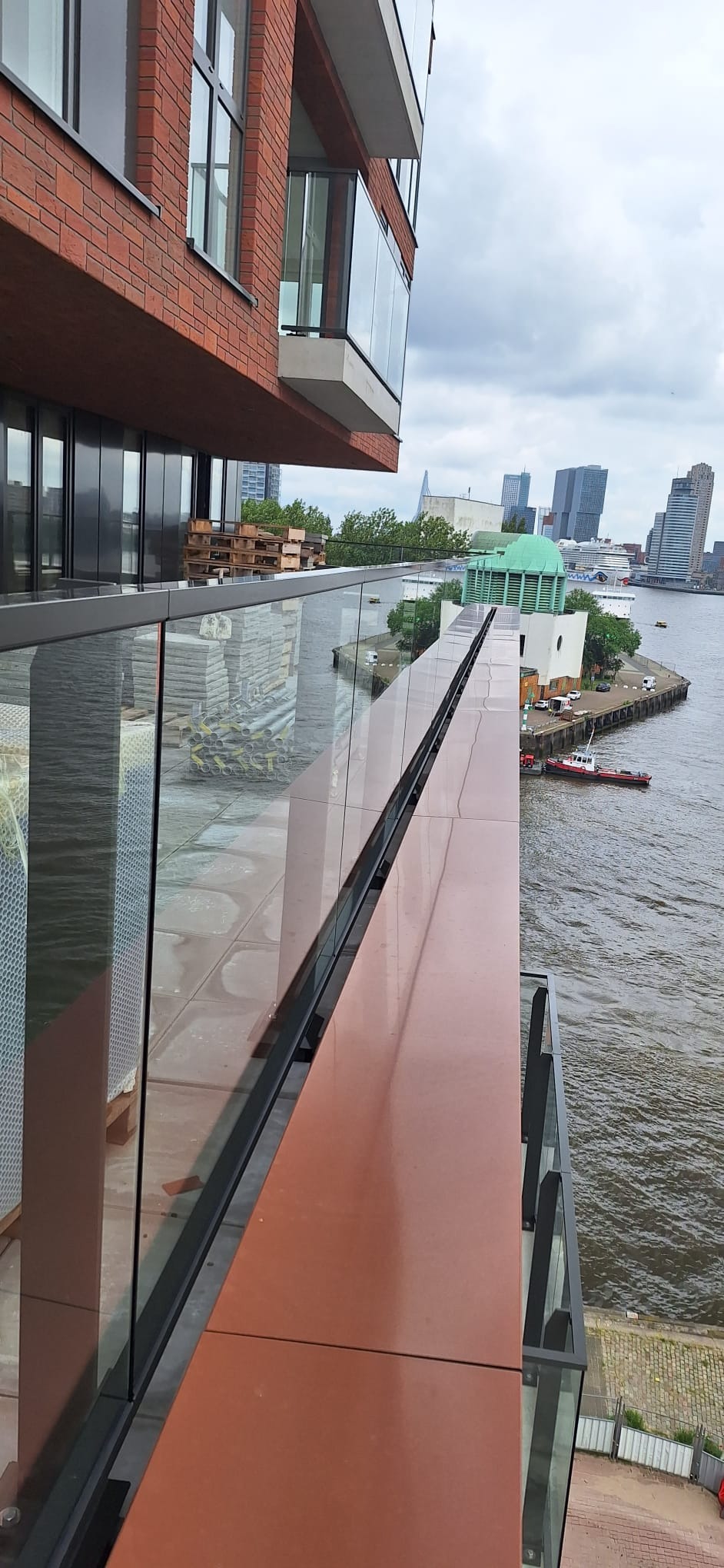 Szklana balustrada balkonowa z widokiem na port i panoramę miasta w tle, brązowe panele elewacyjne.