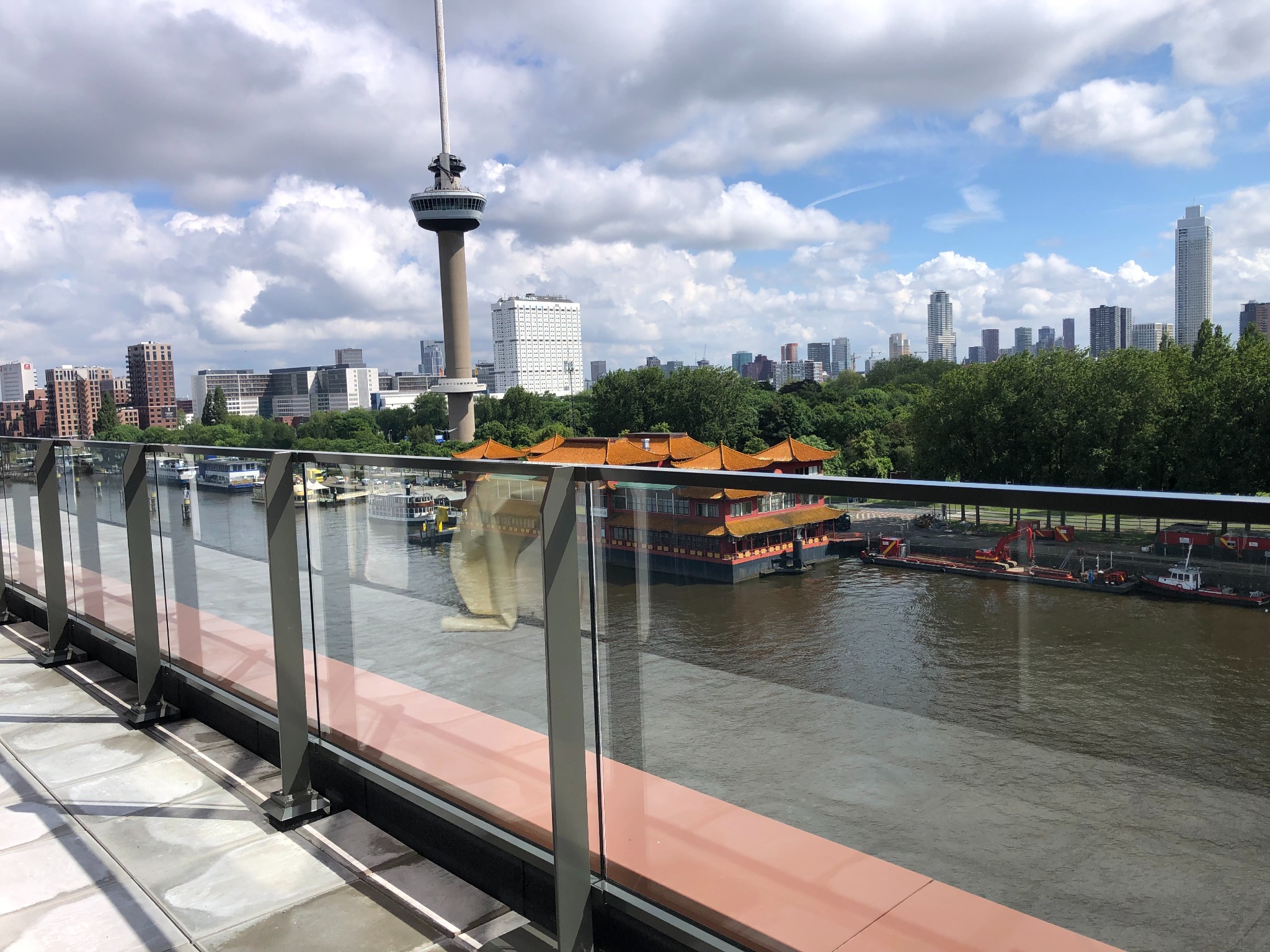 Szklana balustrada na tarasie z widokiem na rzekę, pływający chiński pawilon i panoramę miasta Rotterdam z wieżą Euromast w tle.