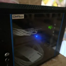 Zamknięta szafa rack z widocznymi kablami i diodami LED, klucz w zamku. Szafa marki CMR Pro, umieszczona na drewnianej powierzchni. Widoczne okno w tle.