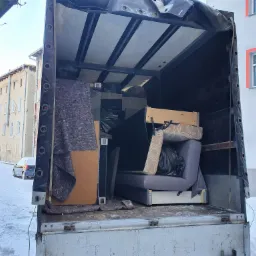 Otwarta paka ciężarówki załadowana meblami i zabezpieczona kocami transportowymi, widok z zewnątrz na zaśnieżone podwórze i budynki.