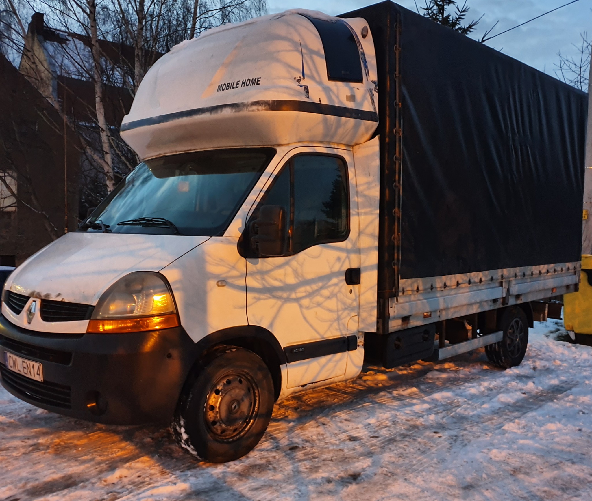 Biały samochód dostawczy Renault Master z czarną plandeką na skrzyni ładunkowej, zaparkowany na śniegu, z napisem 'MOBILE HOME' na kabinie.