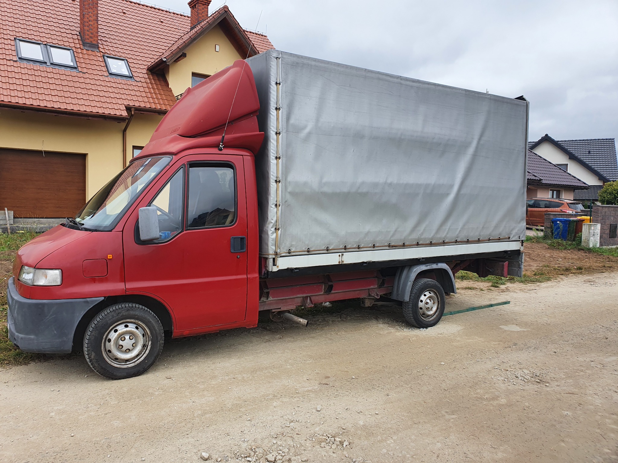 Czerwony samochód dostawczy z szarą plandeką na tle domu z czerwoną dachówką, stojący na piaszczystej drodze.