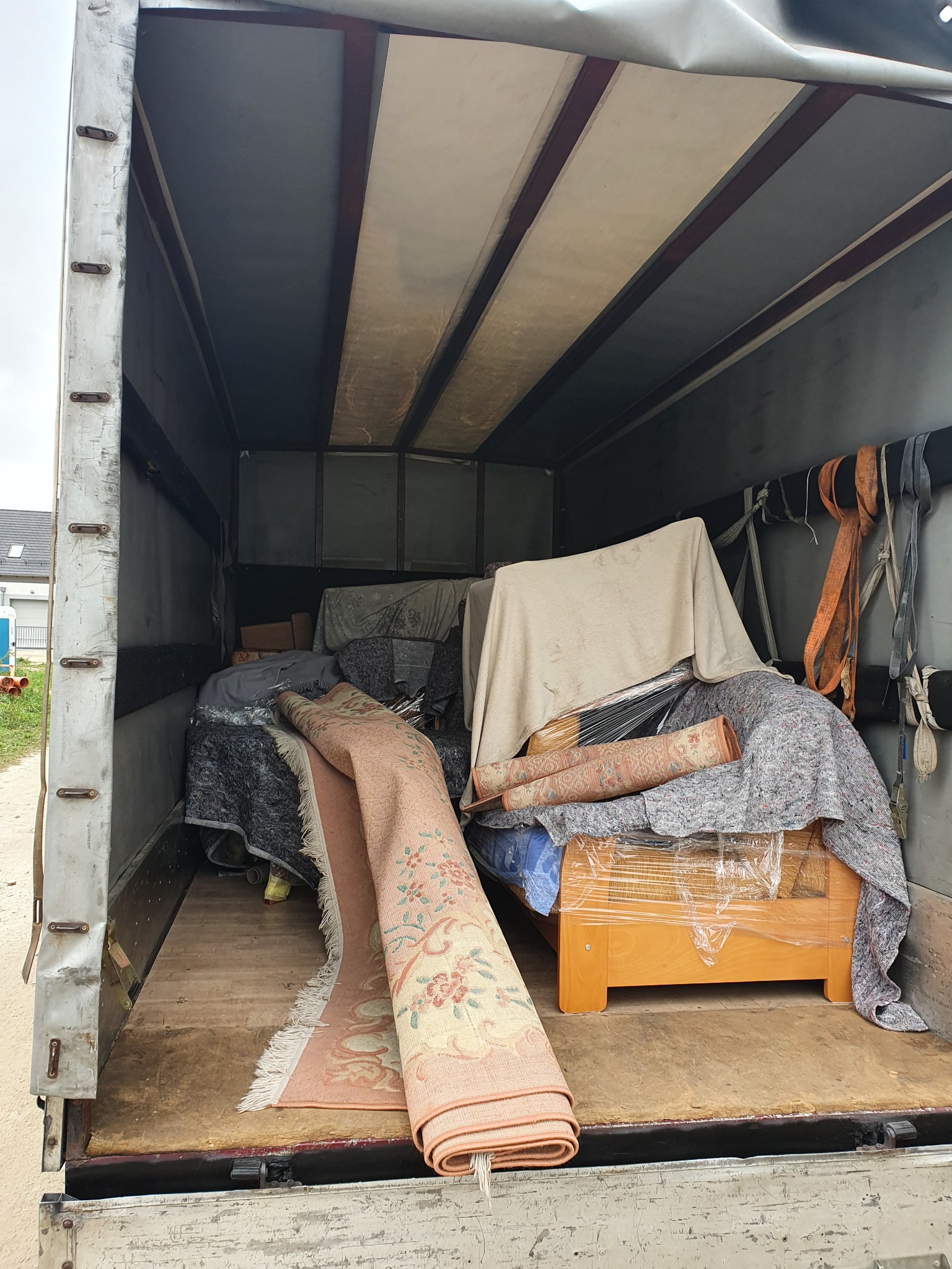Wnętrze paki transportowej wypełnione meblami i dywanami zabezpieczonymi folią i kocami, widoczne pasy mocujące.