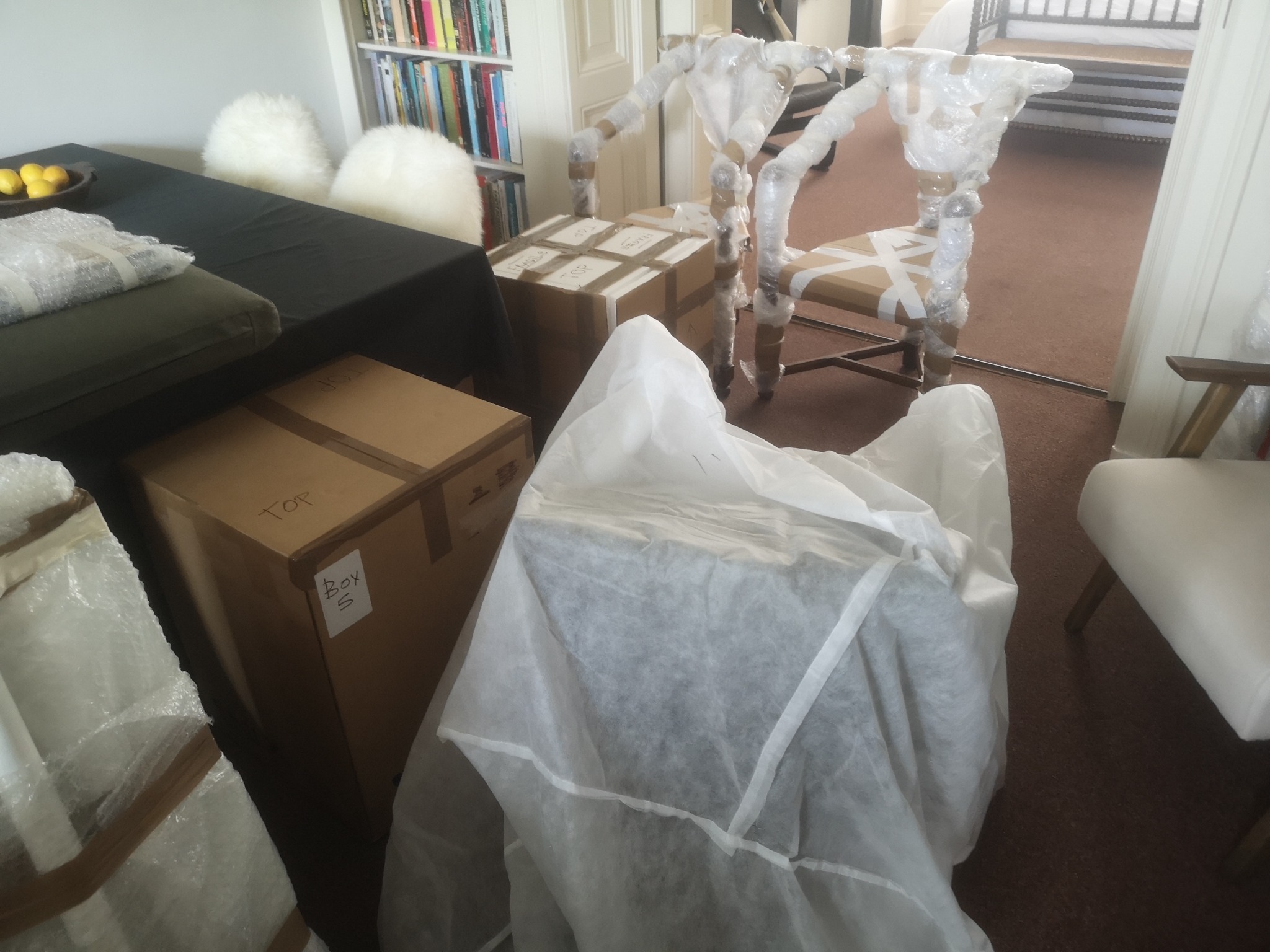 Przygotowane do transportu: meble i kartony spakowane w folię i taśmę, widoczny napis 'TOP' na kartonie i regał z książkami w tle.