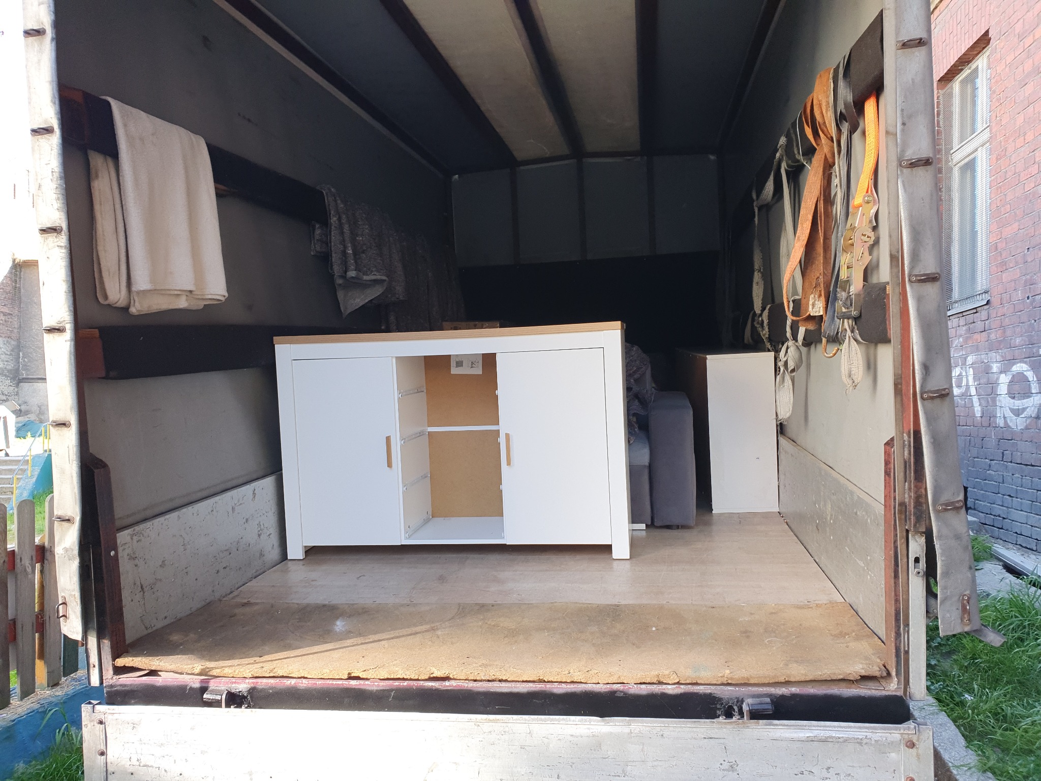 Wnętrze brudnej paki samochodu dostawczego z meblami: biała szafka z otwartymi drzwiczkami, szara sofa i biała szafka po prawej stronie, zabezpieczone pasami transportowymi.