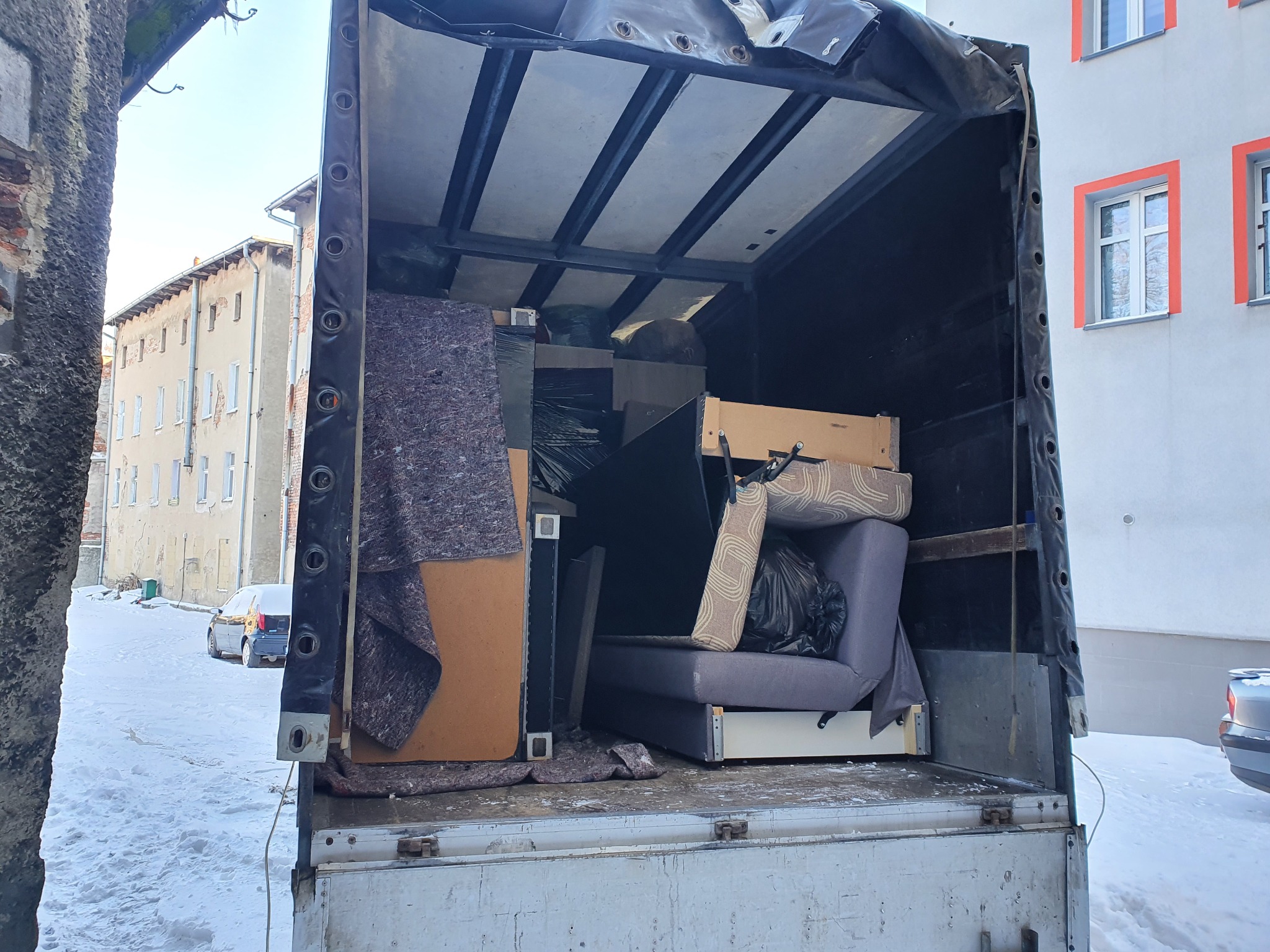 Otwarta paka ciężarówki załadowana meblami i zabezpieczona kocami transportowymi, widok z zewnątrz na zaśnieżone podwórze i budynki.