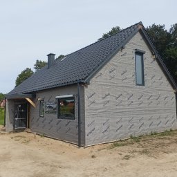 MARDAN DOM SPÓŁKA Z OGRANICZONĄ ODPOWIEDZIALNOŚCIĄ - Surowy stan domu z czarną dachówką, ocieplony membraną Strotex, z widocznymi oknami i przygotowanym wejściem, na piaszczystym terenie.