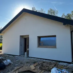 Surowy stan elewacji parterowego domu jednorodzinnego w Gołkowicach Dolnych z ciemną stolarką okienną i czarnym dachem, widoczne ślady budowy i resztki materiałów budowlanych.