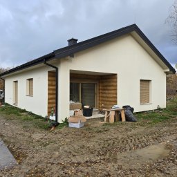 MARDAN DOM SPÓŁKA Z OGRANICZONĄ ODPOWIEDZIALNOŚCIĄ - Nowoczesny, parterowy dom z jasną elewacją i drewnianym wykończeniem, widoczny w pochmurny dzień, z elementami budowy w otoczeniu, takimi jak skrzynki i stół roboczy.