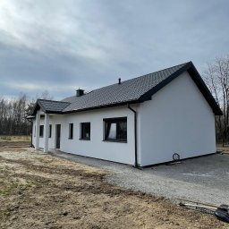 MARDAN DOM - Gotowy, parterowy dom z białymi ścianami i ciemną dachówką, widoczny z boku. Na pierwszym planie ziemia i żwir, w tle drzewa bez liści. Pochmurne niebo.