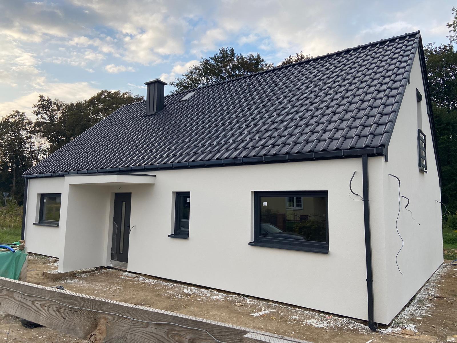 Nowoczesny, parterowy dom z ciemną dachówką i minimalistycznymi oknami, w trakcie wykończenia elewacji, widoczne przewody na ścianie.
