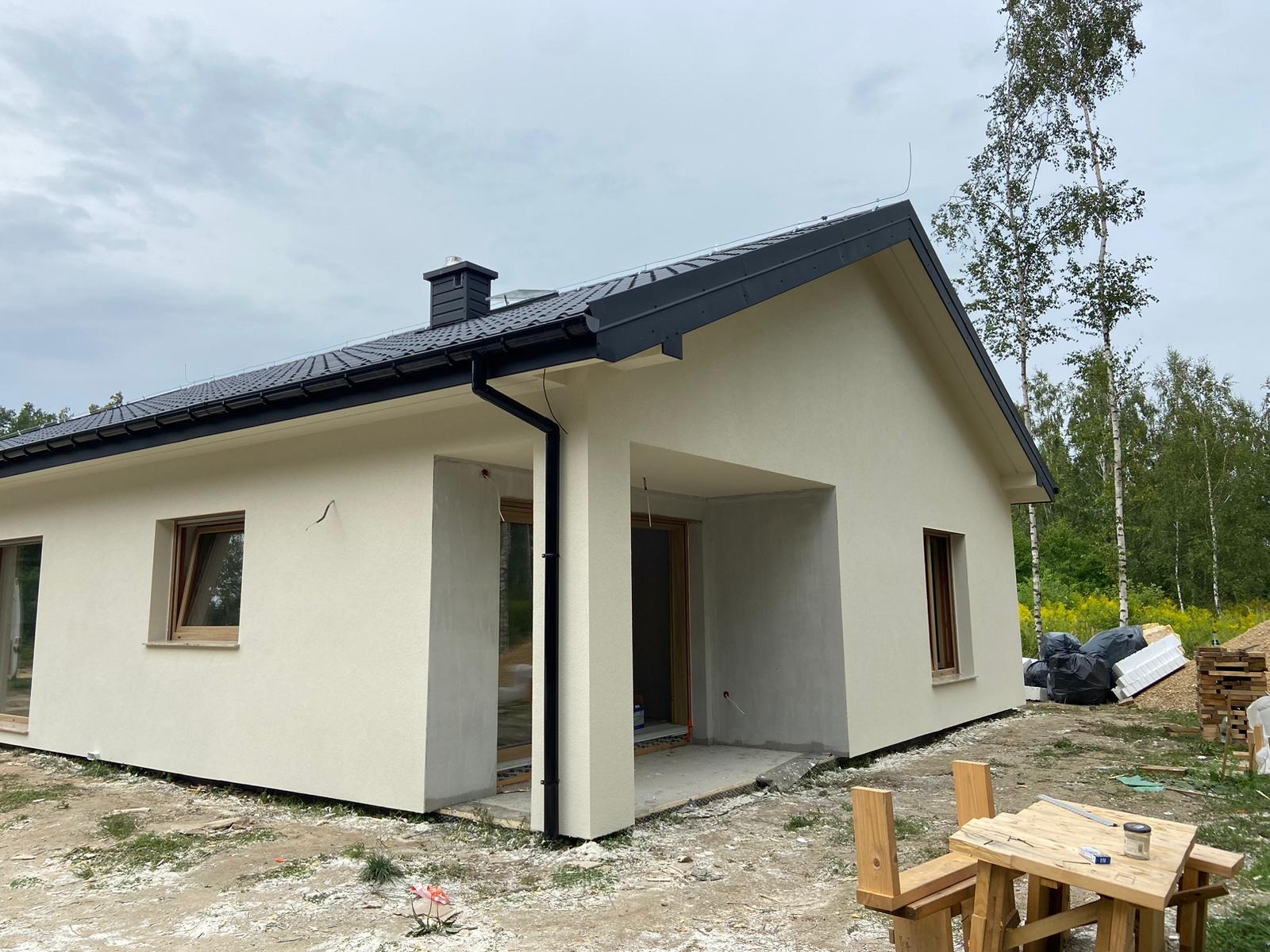 Nowoczesny parterowy dom z dachem dwuspadowym i czarną dachówką, widok z boku, z elementami wykończeniowymi na zewnątrz i drewnianym stolikiem warsztatowym z narzędziami na pierwszym planie.