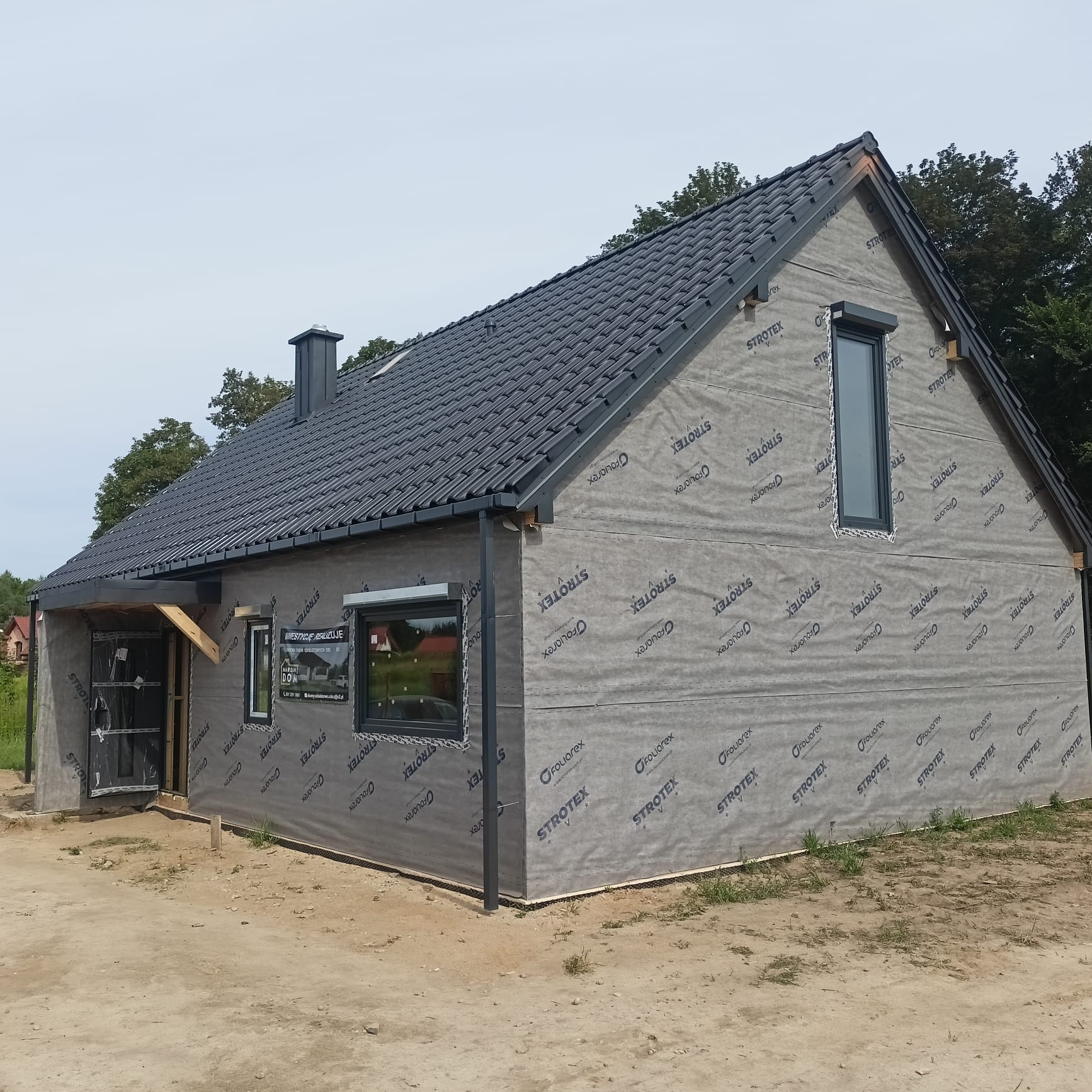 Surowy stan domu z czarną dachówką, ocieplony membraną Strotex, z widocznymi oknami i przygotowanym wejściem, na piaszczystym terenie.