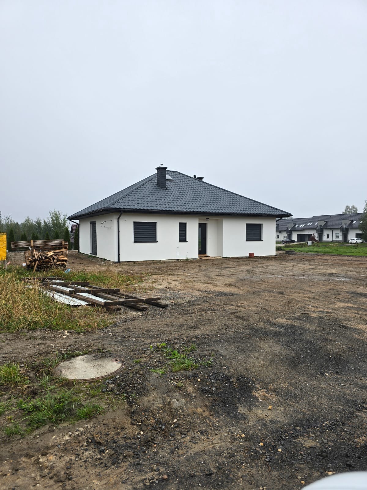 Nowoczesny, parterowy dom szkieletowy z ciemnoszarą dachówką, białymi ścianami i czarnymi roletami zewnętrznymi, widoczny na działce z przygotowanym terenem pod trawnik, w tle inne domy.