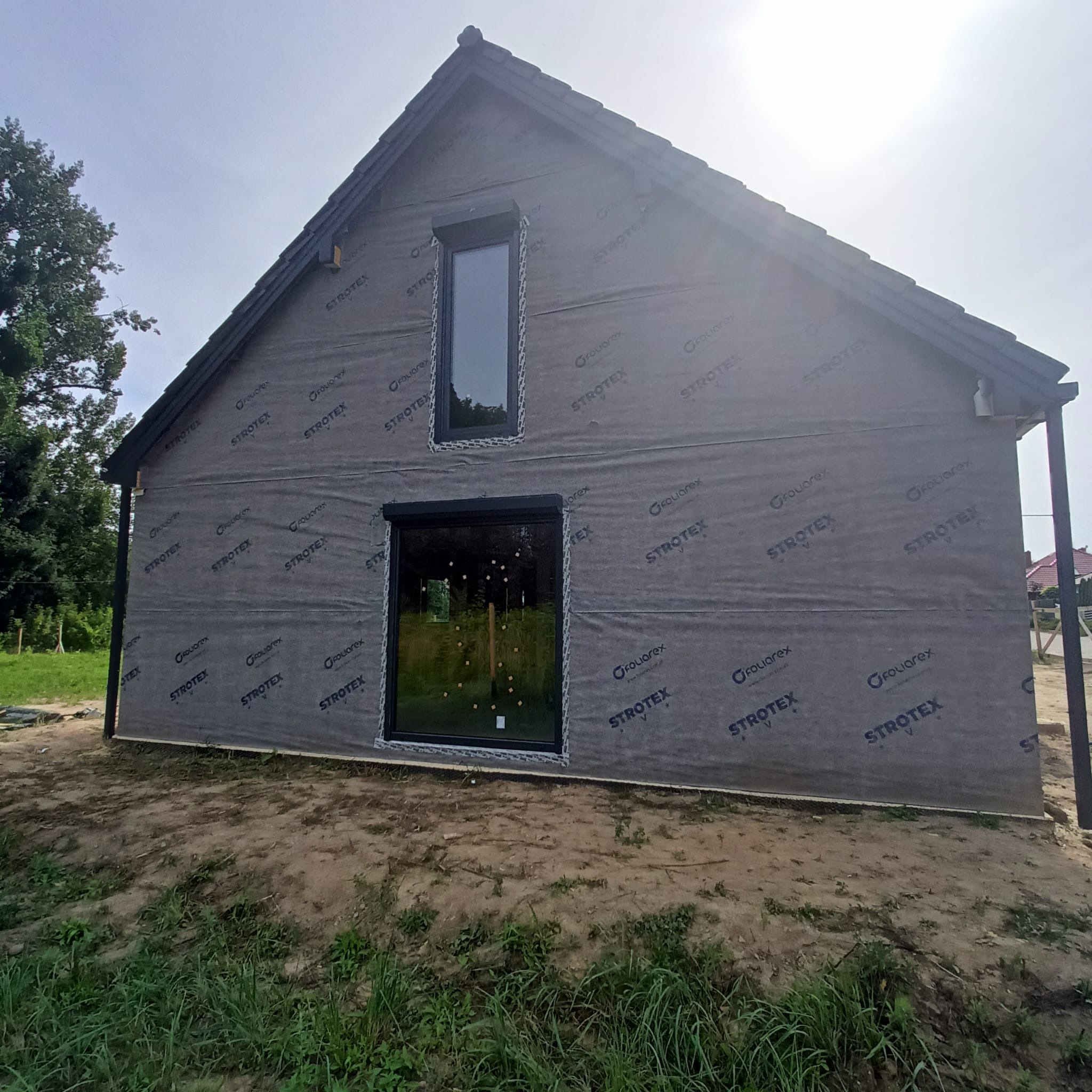 Surowy stan elewacji domu podczas budowy, pokrytej membraną wiatroizolacyjną Strotex z widocznymi oknami z czarnymi ramami i roletami zewnętrznymi, na tle zieleni.