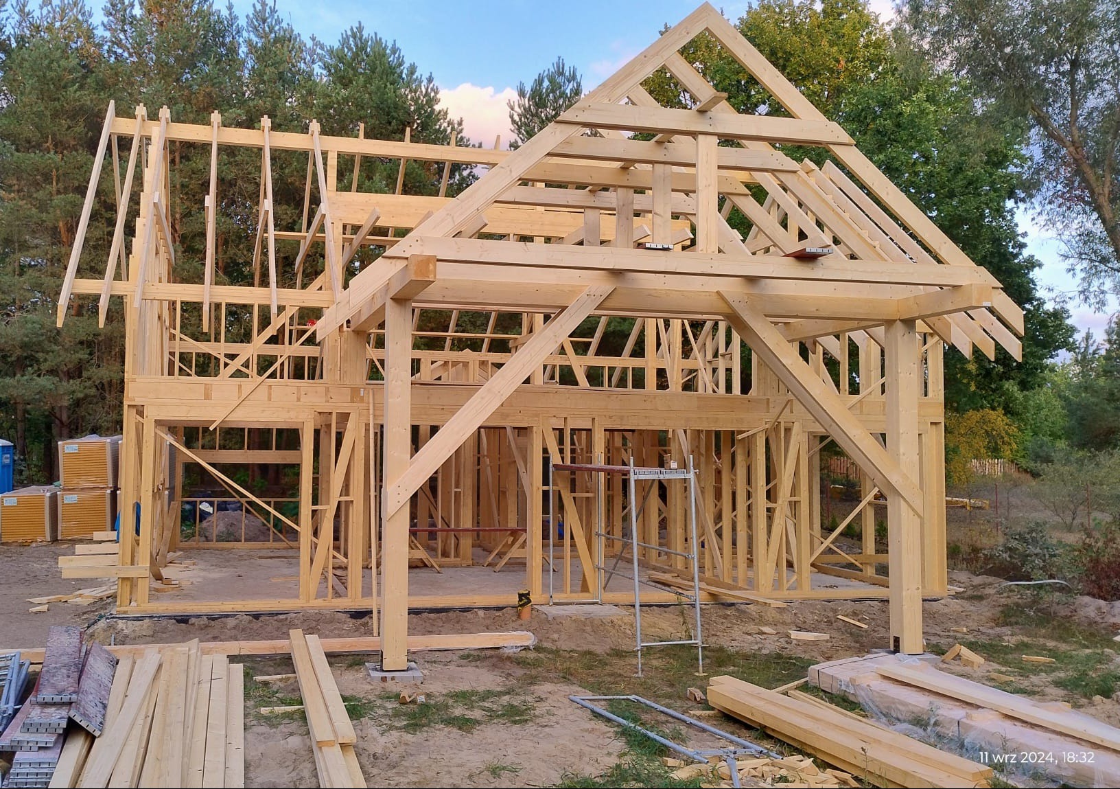 Konstrukcja drewniana domu w trakcie budowy, widoczne elementy szkieletu, rusztowanie i zapas desek na ziemi, w tle drzewa.