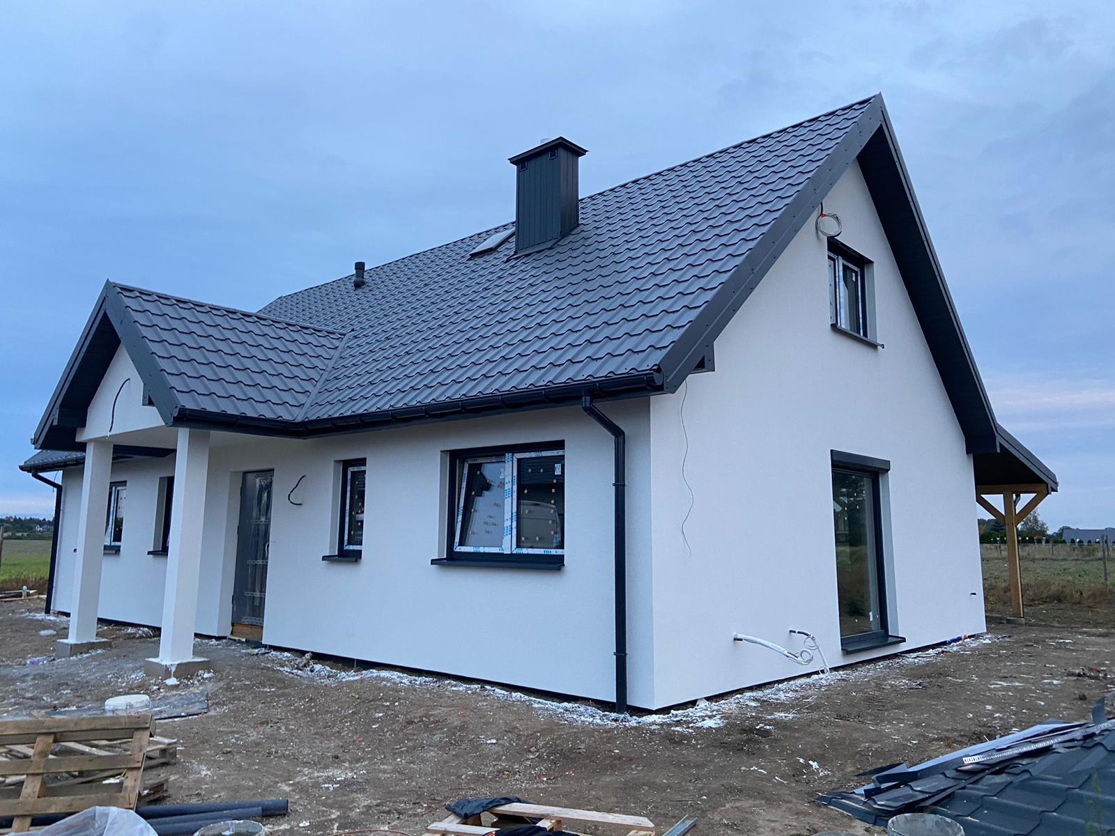 Niedokończony biały dom z szarą dachówką i czarnymi ramami okiennymi, widoczne przewody na elewacji i materiały budowlane na ziemi.