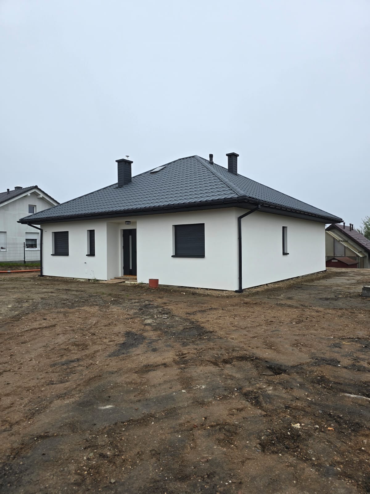 Nowoczesny, parterowy budynek z białymi ścianami i ciemną dachówką, czarnymi oknami z roletami zewnętrznymi oraz dwoma kominami, otoczony ziemią bez trawy.