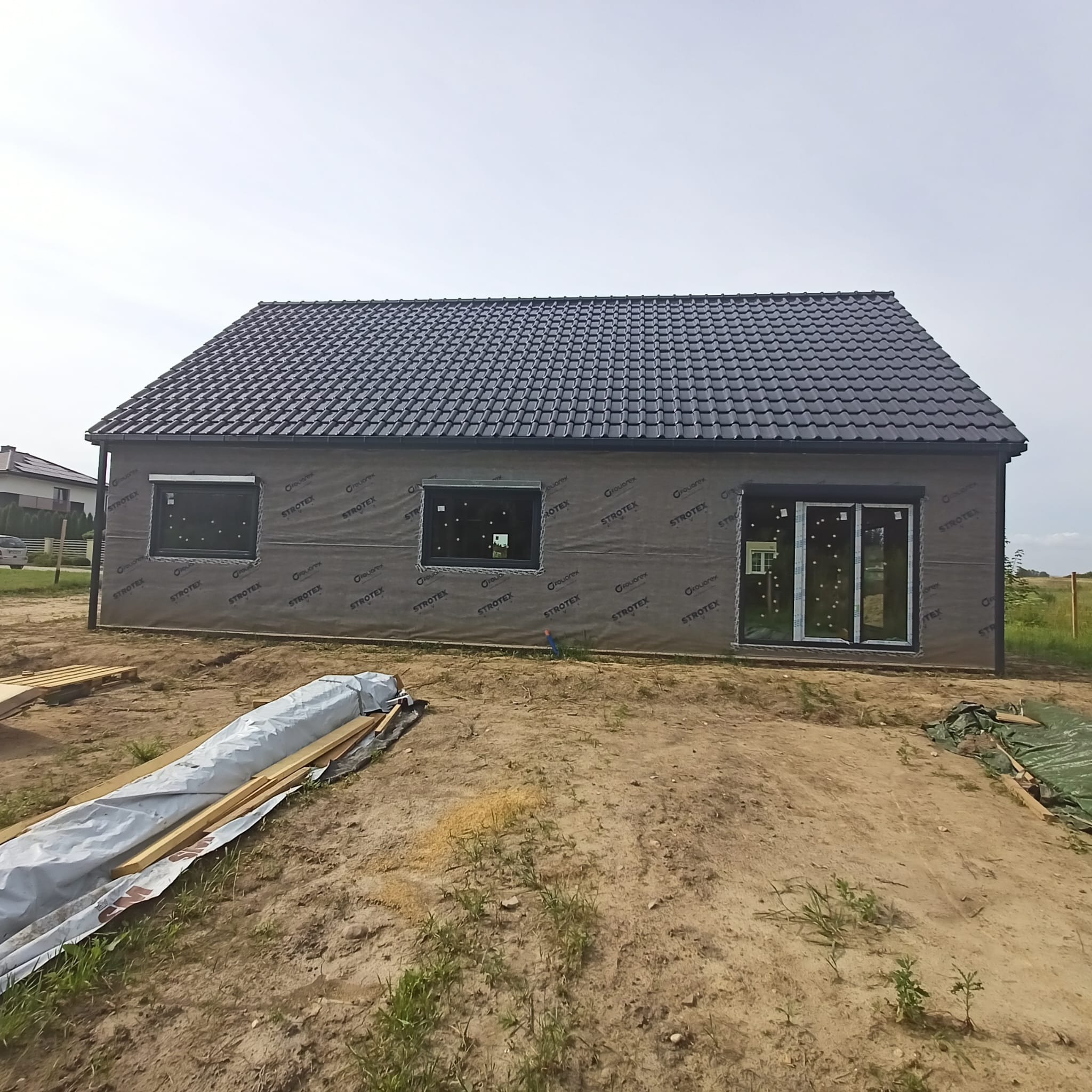 Surowy stan domu z ciemną dachówką, pokrytego folią izolacyjną z nadrukiem, z oknami z roletami zewnętrznymi, na tle piaszczystego placu budowy z materiałami budowlanymi.
