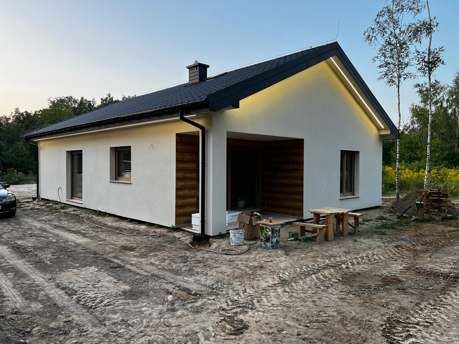 Nowoczesny, parterowy budynek z elewacją w kolorze złamanej bieli, podświetlony listwą LED pod okapem, z drewnianym wykończeniem wnęki wejściowej oraz czarnym dachem z dachówki, widoczny wczesnym...