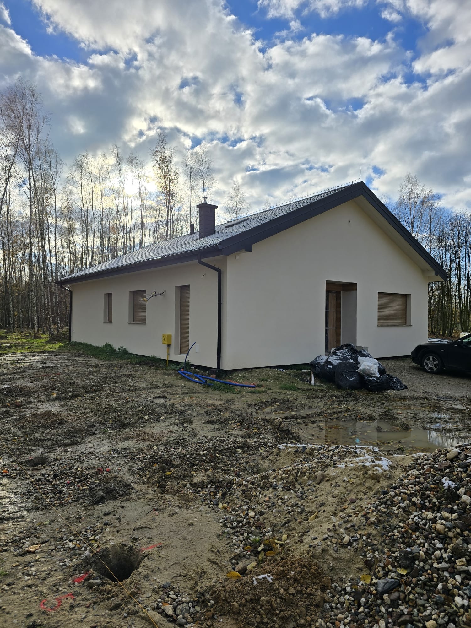 Niedokończony parterowy budynek z jasną elewacją i ciemnym dachem na działce w trakcie przygotowania, widoczne wykopane doły i materiały budowlane, w tle drzewa bez liści i pochmurne niebo.