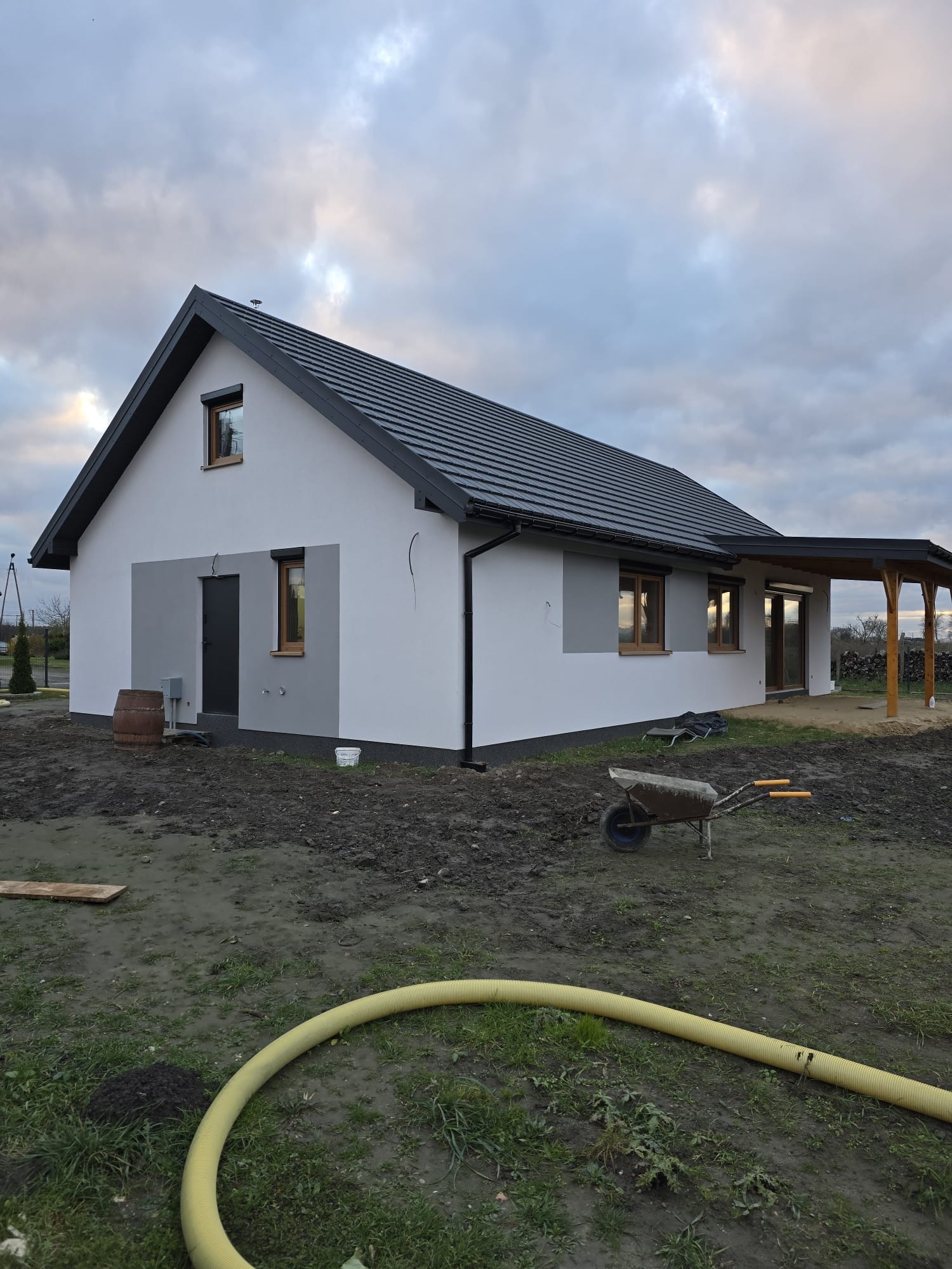 Nowoczesny, parterowy budynek z dachem dwuspadowym i drewnianą pergolą, widoczny w pochmurny dzień, z żółtym wężem ogrodowym i taczką na pierwszym planie.