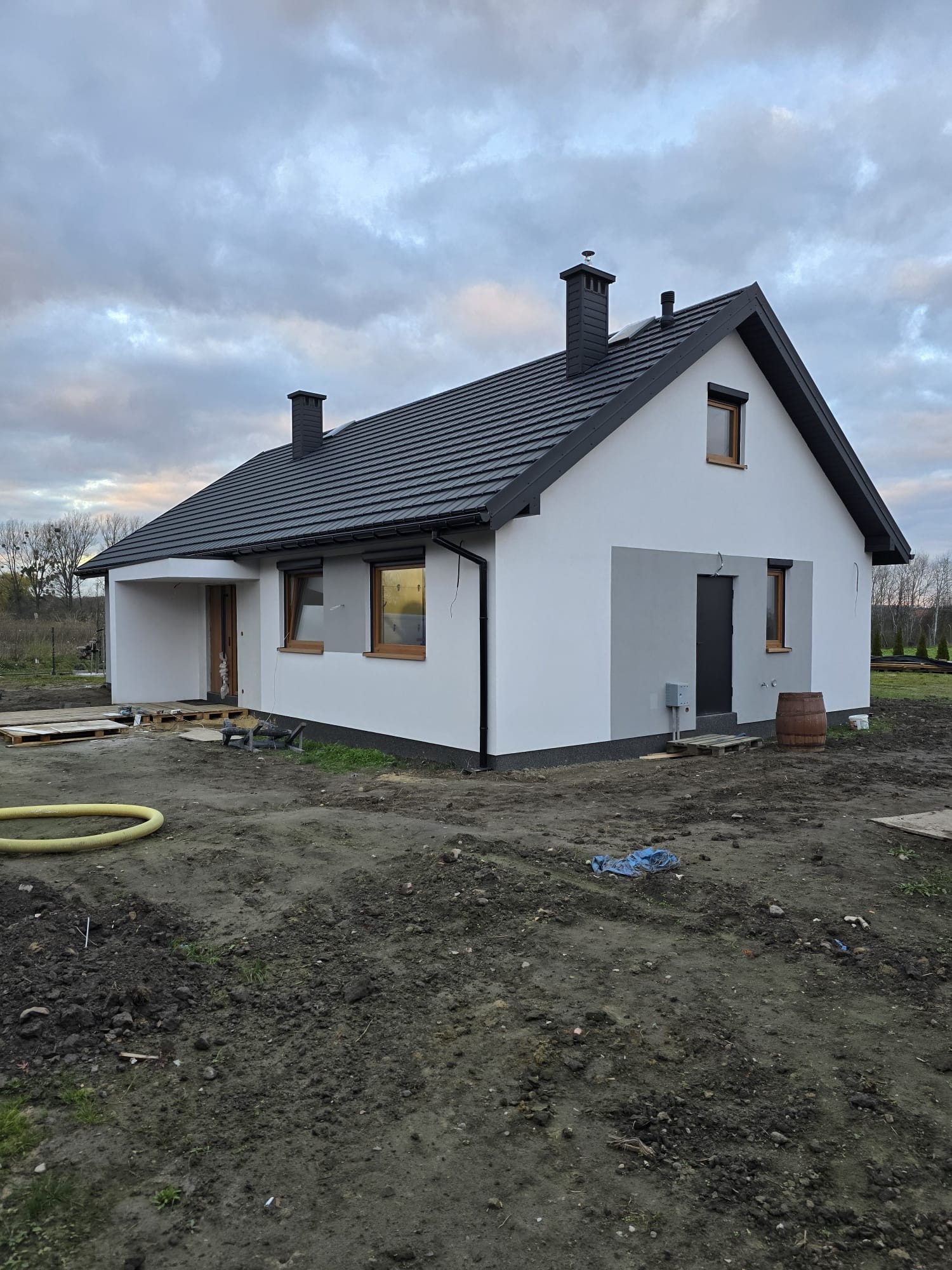 Niedokończony dom szkieletowy z ciemną dachówką i białymi ścianami, widoczne elementy budowlane na posesji, pochmurne niebo.