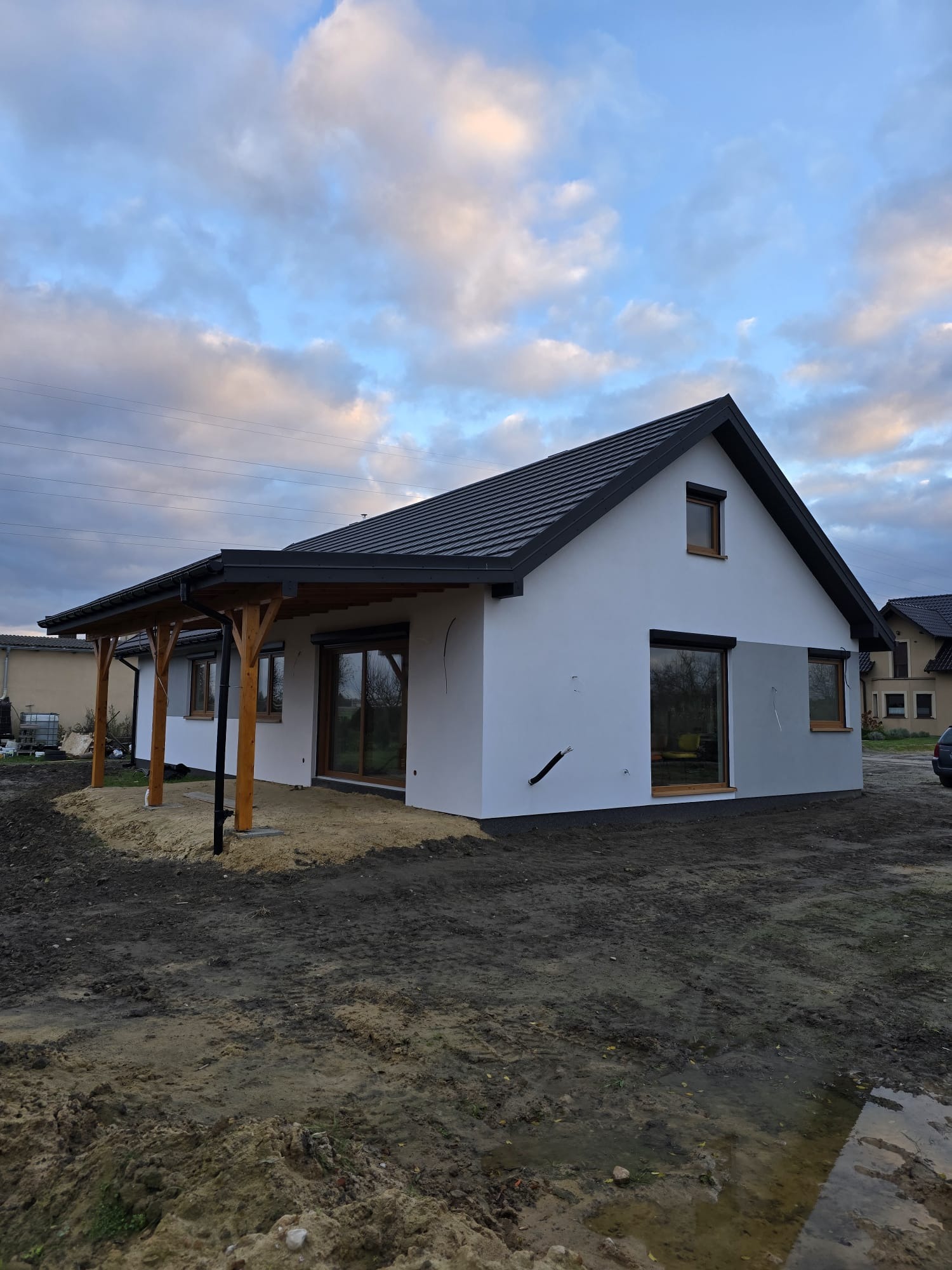 Surowy stan elewacji jednokondygnacyjnego domu z drewnianą konstrukcją zadaszenia tarasu i czarną dachówką, widok z zewnątrz na tle pochmurnego nieba.