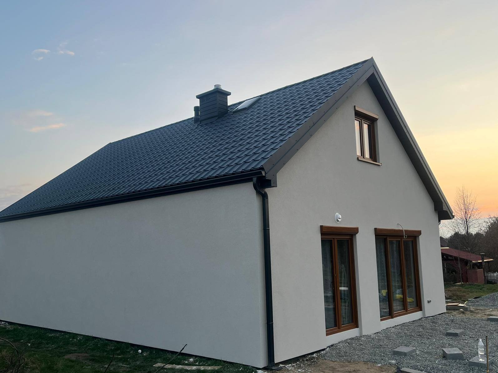 Gotowy dom z ciemną dachówką i brązowymi oknami, widziany z boku na tle zachodzącego słońca. Widoczny komin i rynny. Elewacja w jasnym odcieniu szarości.