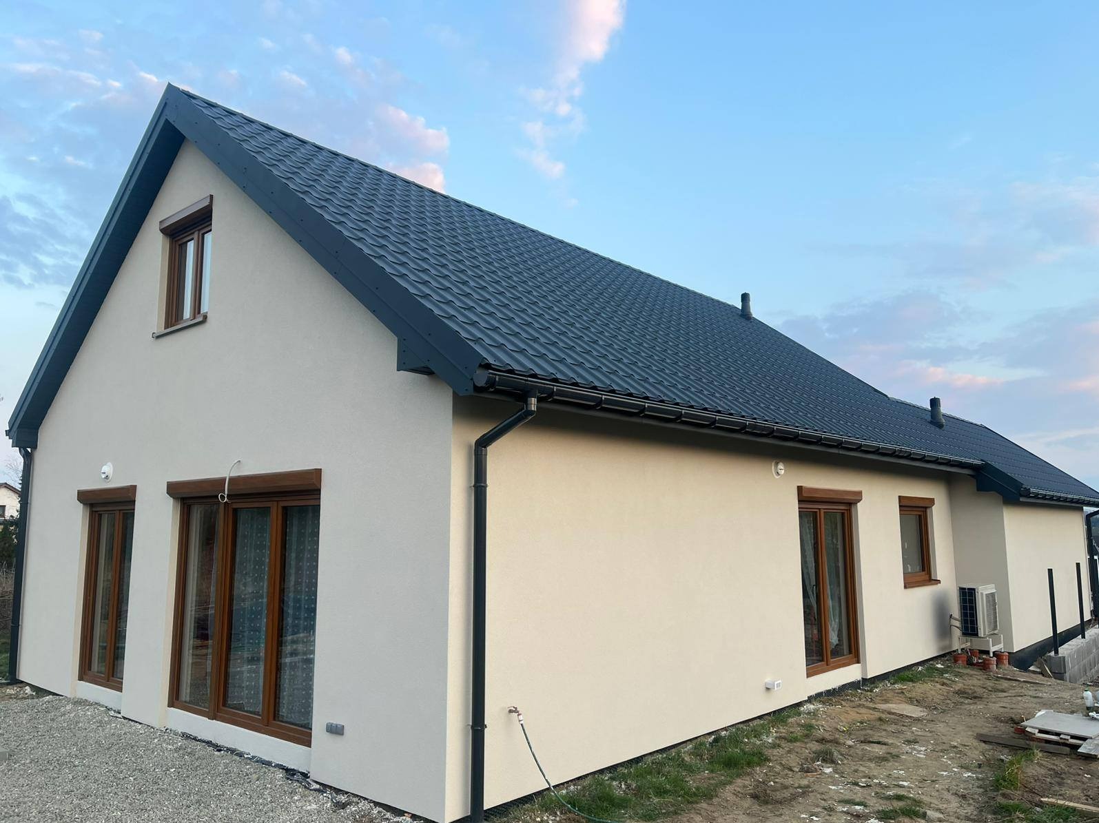 Gotowy dom z ciemną dachówką i jasną elewacją, brązowe okna z roletami, widok z boku, trawa i gruz wokół. Nowoczesny styl, świeżo po budowie.