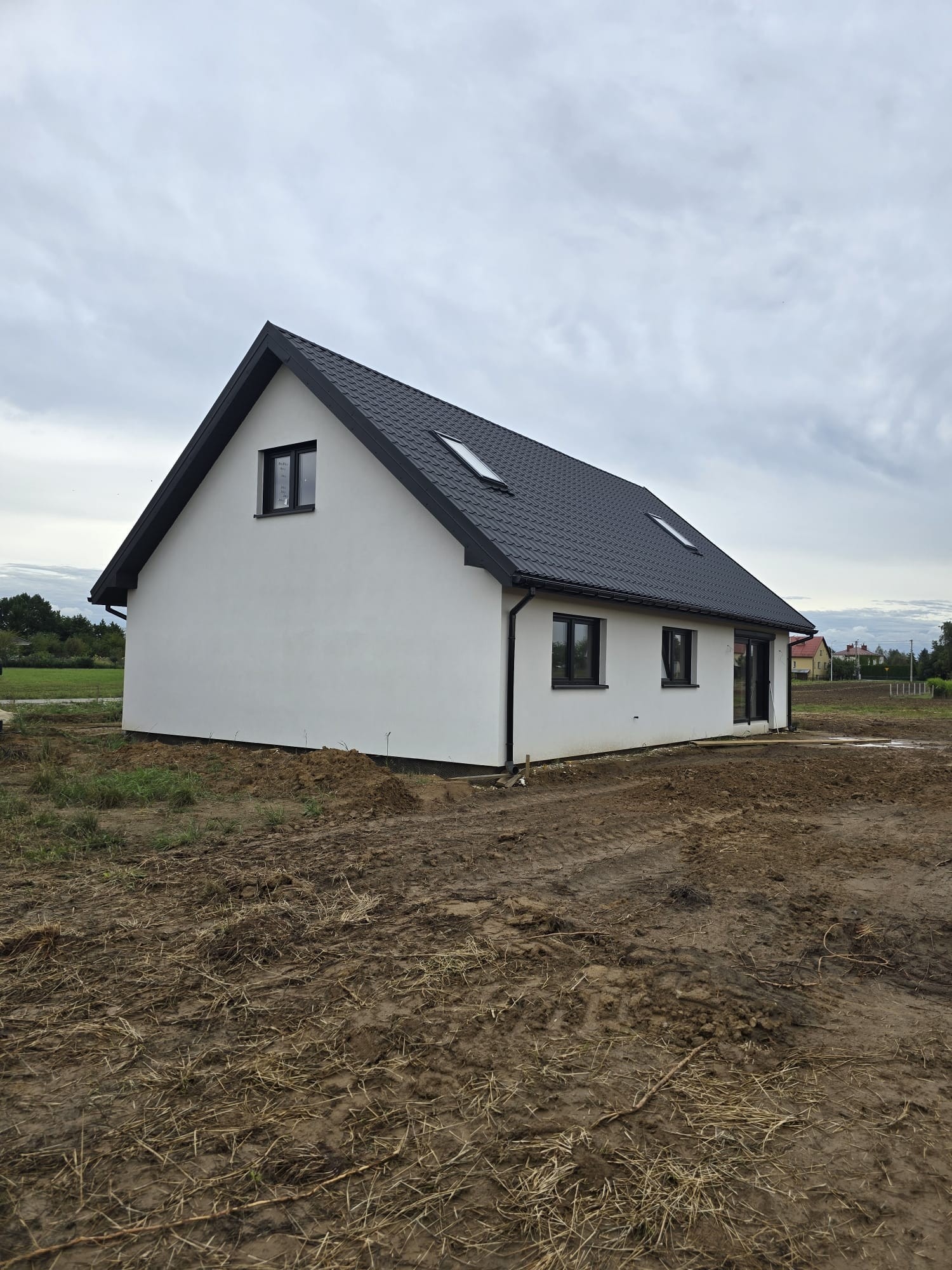 Biały dom z czarnym dachem i oknami, w trakcie budowy, otoczony ziemią. Widoczne ślady opon. Pochmurne niebo w tle. Minimalistyczny styl.