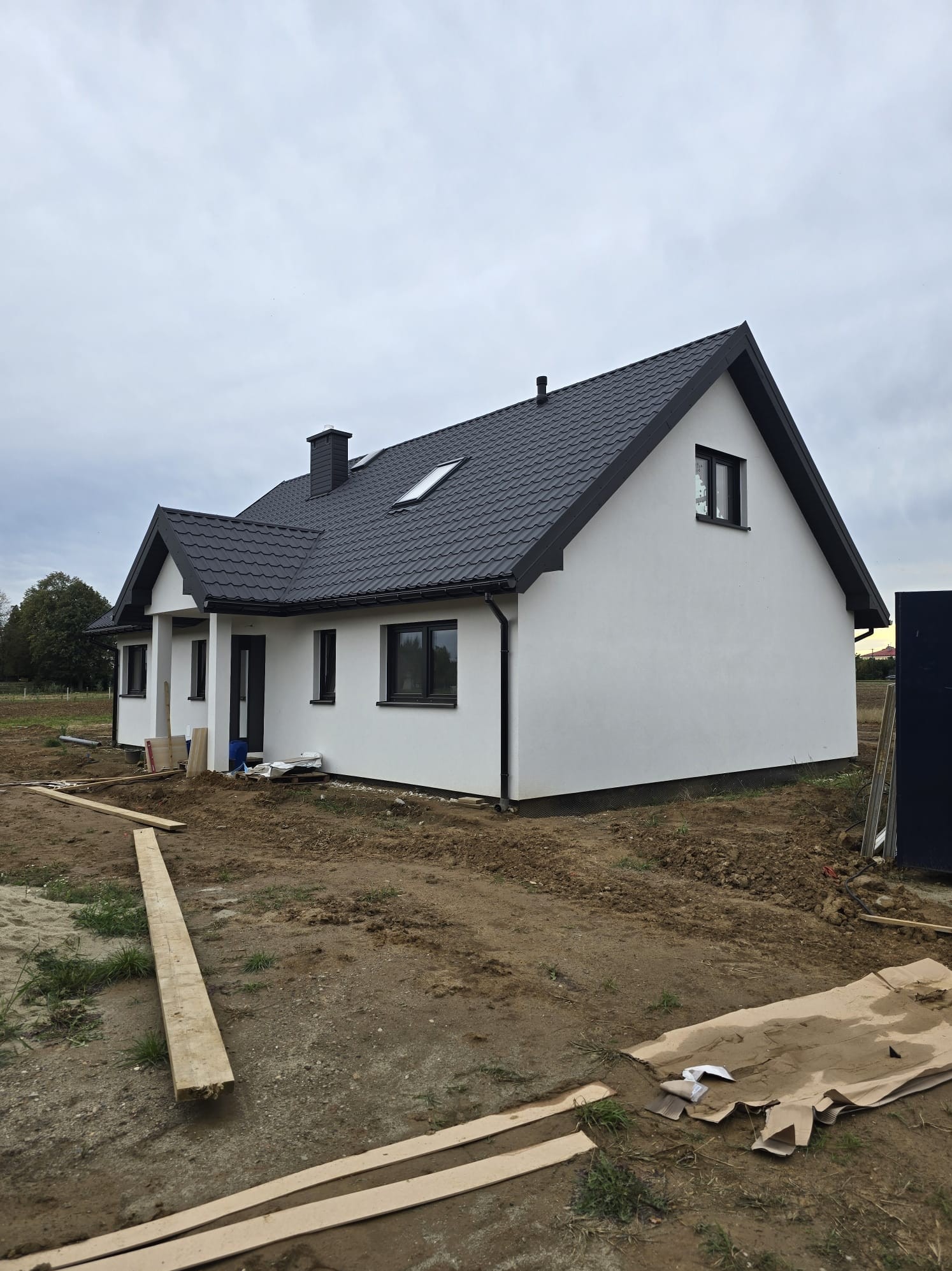 Biały dom z czarnym dachem z blachy trapezowej i kominem, widok z zewnątrz na plac budowy z drewnianymi elementami. Pochmurne niebo w tle.