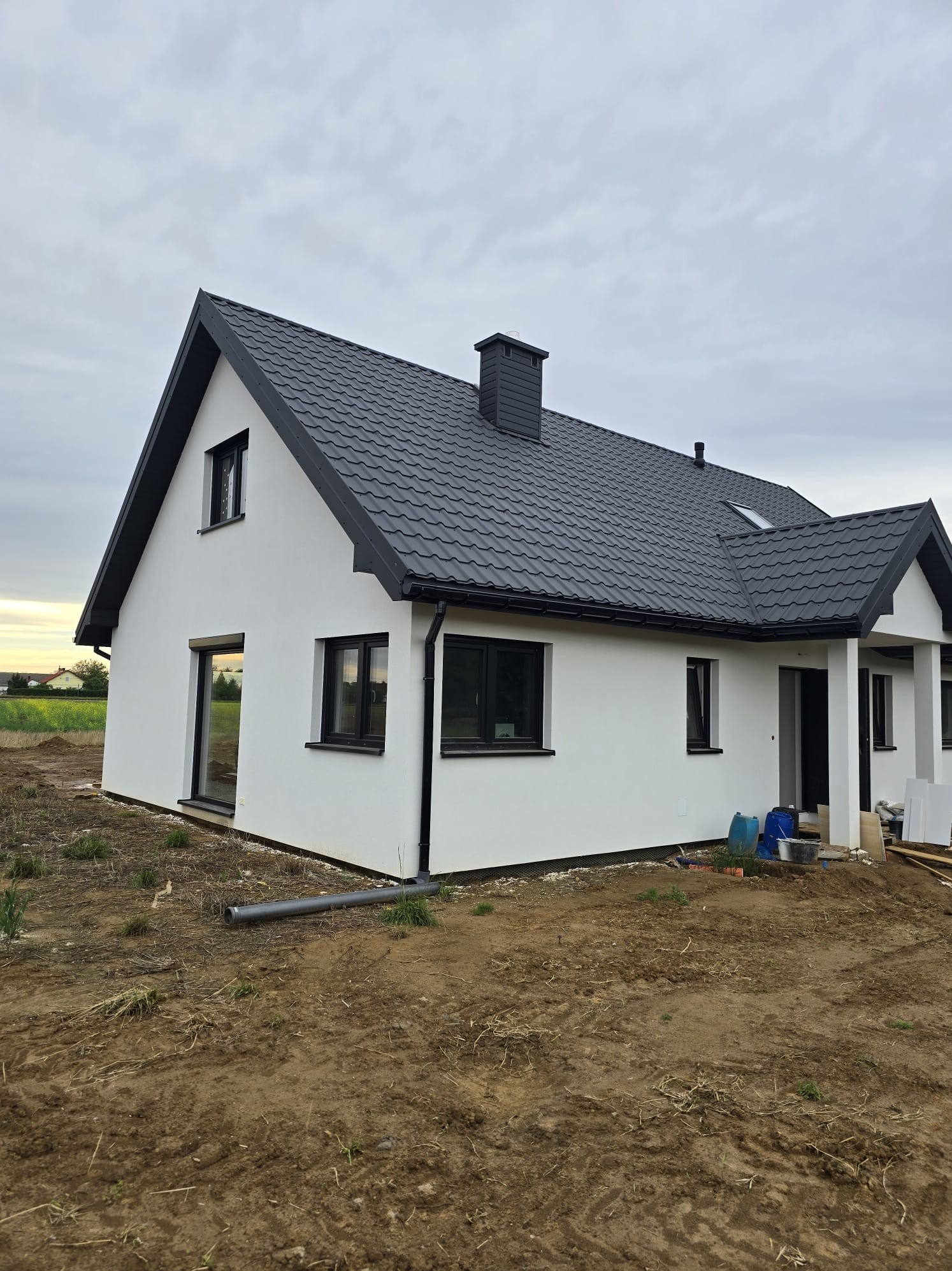 Biały dom z ciemną dachówką i oknami, widoczny z boku na tle pochmurnego nieba i pola. Na pierwszym planie ziemia z resztkami roślinności i rura odpływowa.