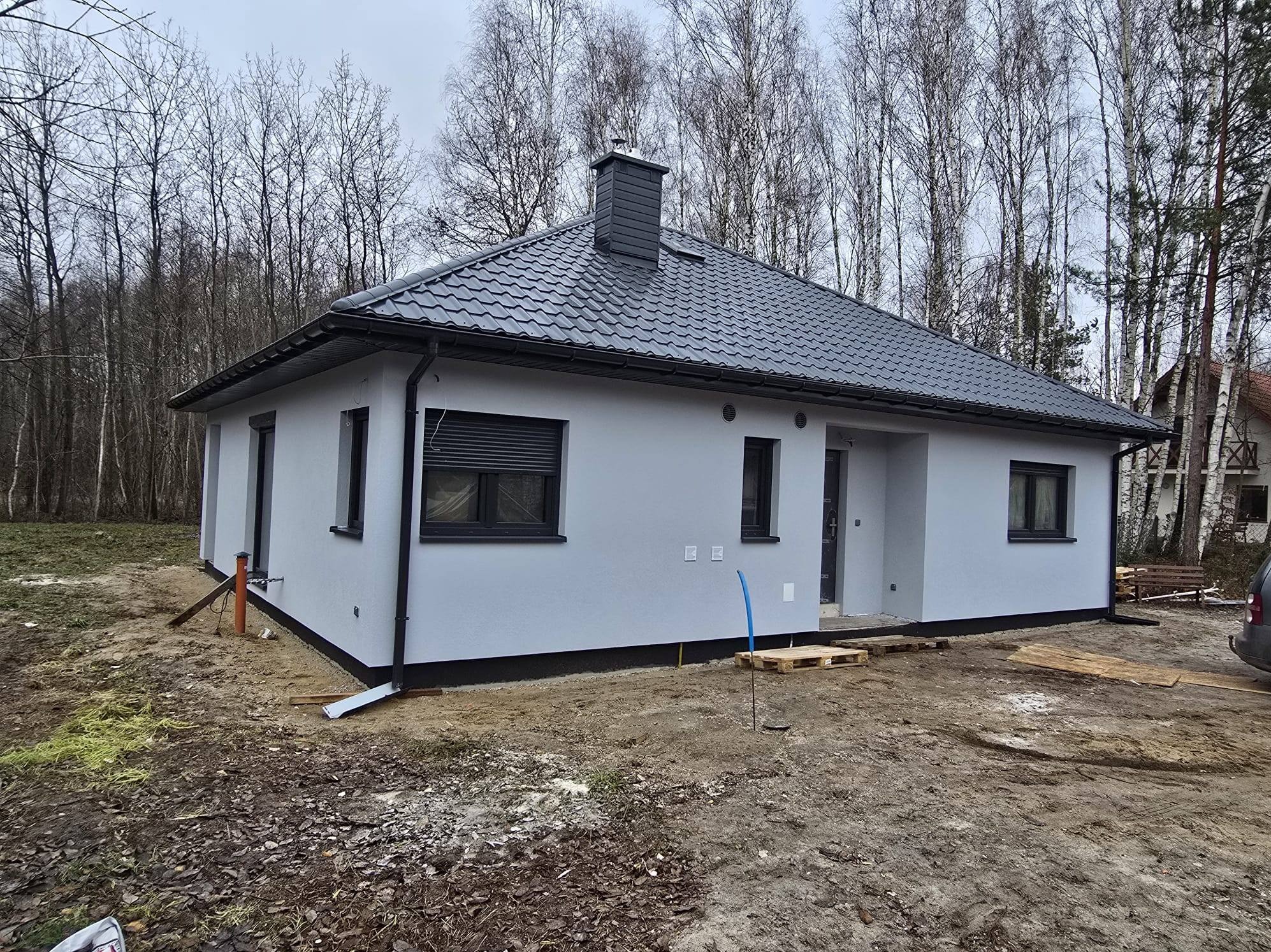 Gotowy, parterowy dom z szarą elewacją i ciemną dachówką na tle drzew. Widoczne detale wykończenia, takie jak rynny i okna z roletami zewnętrznymi.