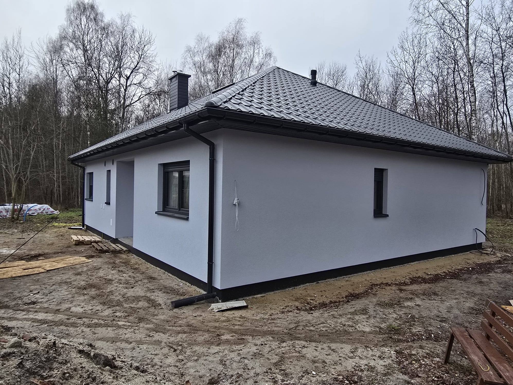 Gotowy, parterowy dom z szarą elewacją i ciemną dachówką, widoczny z boku na tle drzew. Uwagę zwraca minimalistyczny styl i brak wykończenia terenu wokół budynku.